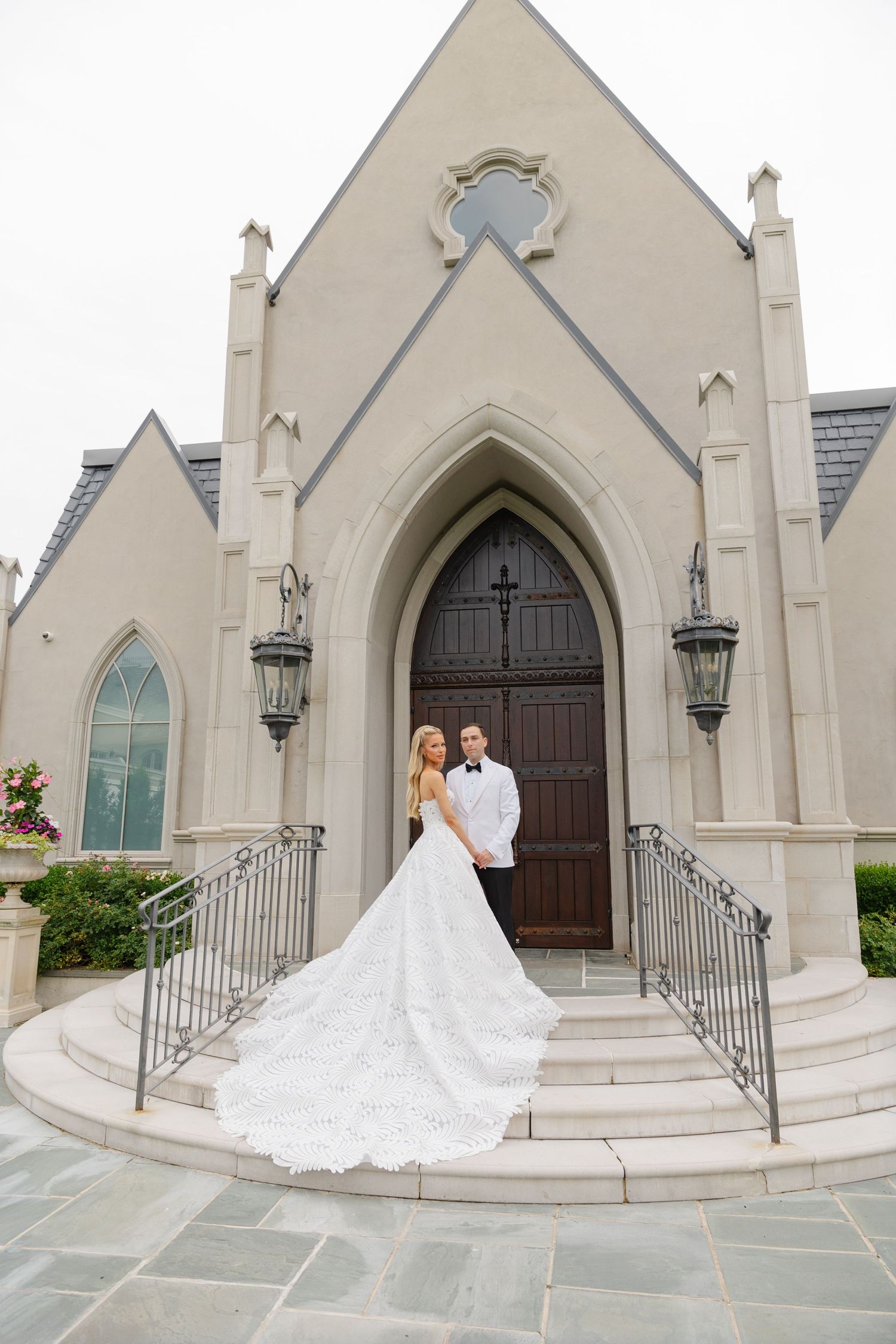 A wedding couple stands on the steps of a Gothic-style white church with arched doorways and decorative railings.