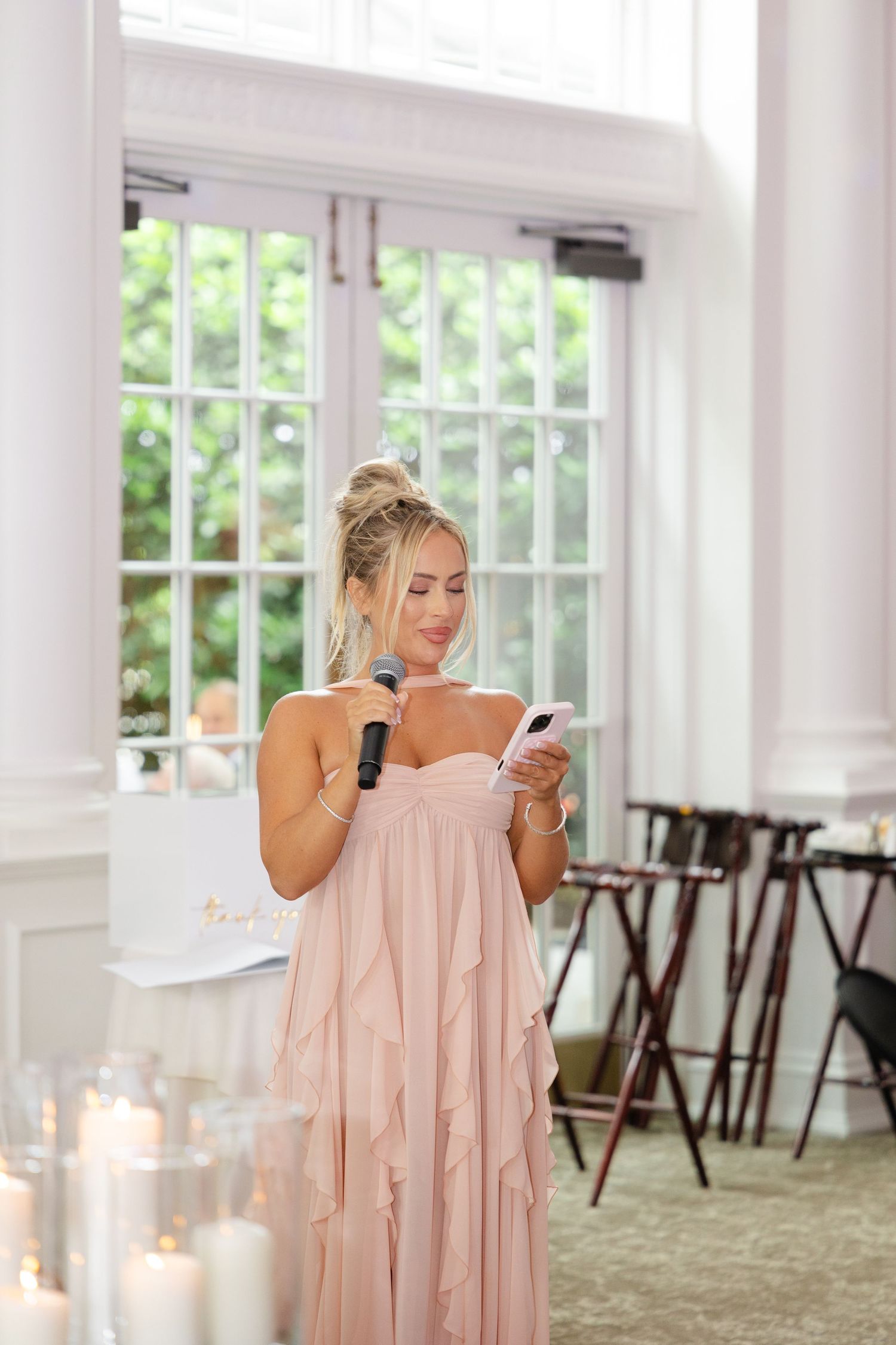 Someone in a flowing blush pink dress checks their phone while standing in an elegant room with candles and windows.