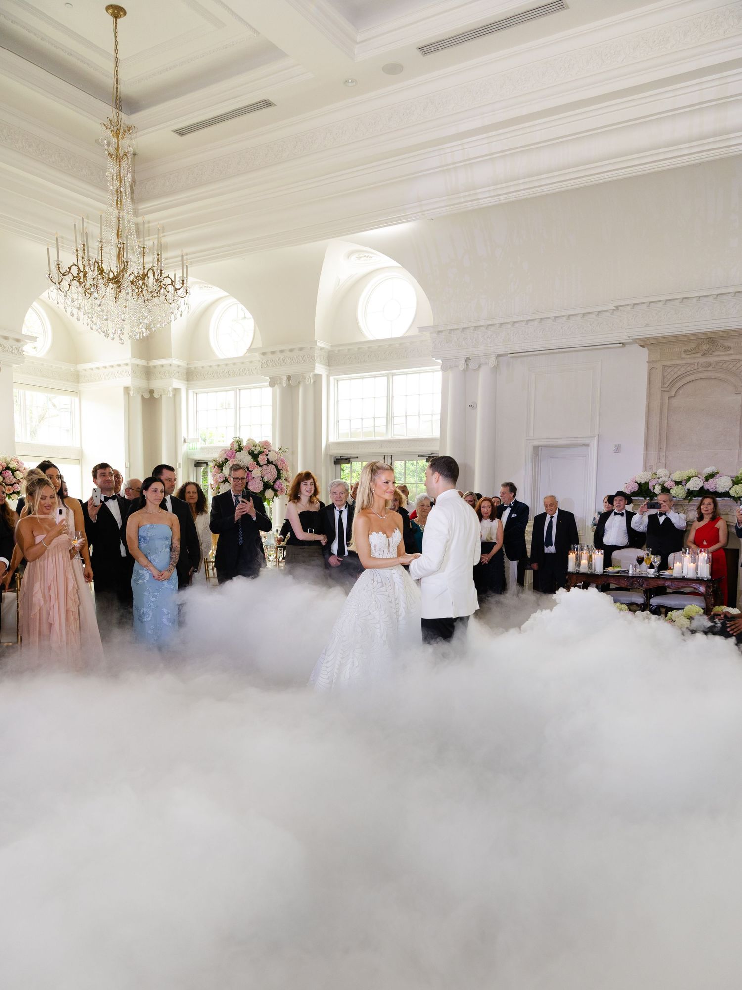 Guests dance on a misty dance floor under elegant chandeliers in a grand white ballroom during a wedding reception.