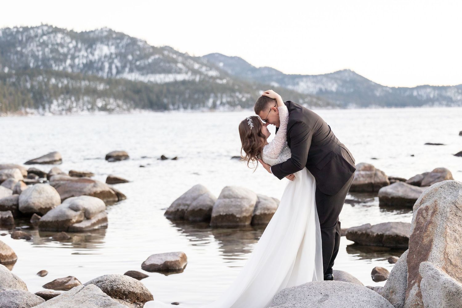 Romantic wedding photos captured on snow-covered rocky beach with mountain landscape.