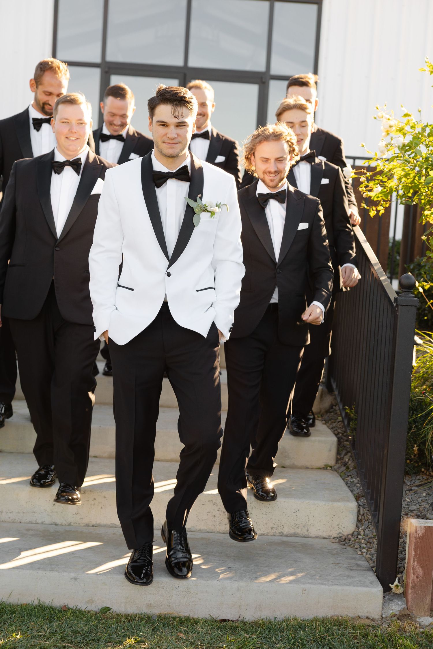 Groomsmen in black and white tuxedos walk together down outdoor steps on a sunny day.