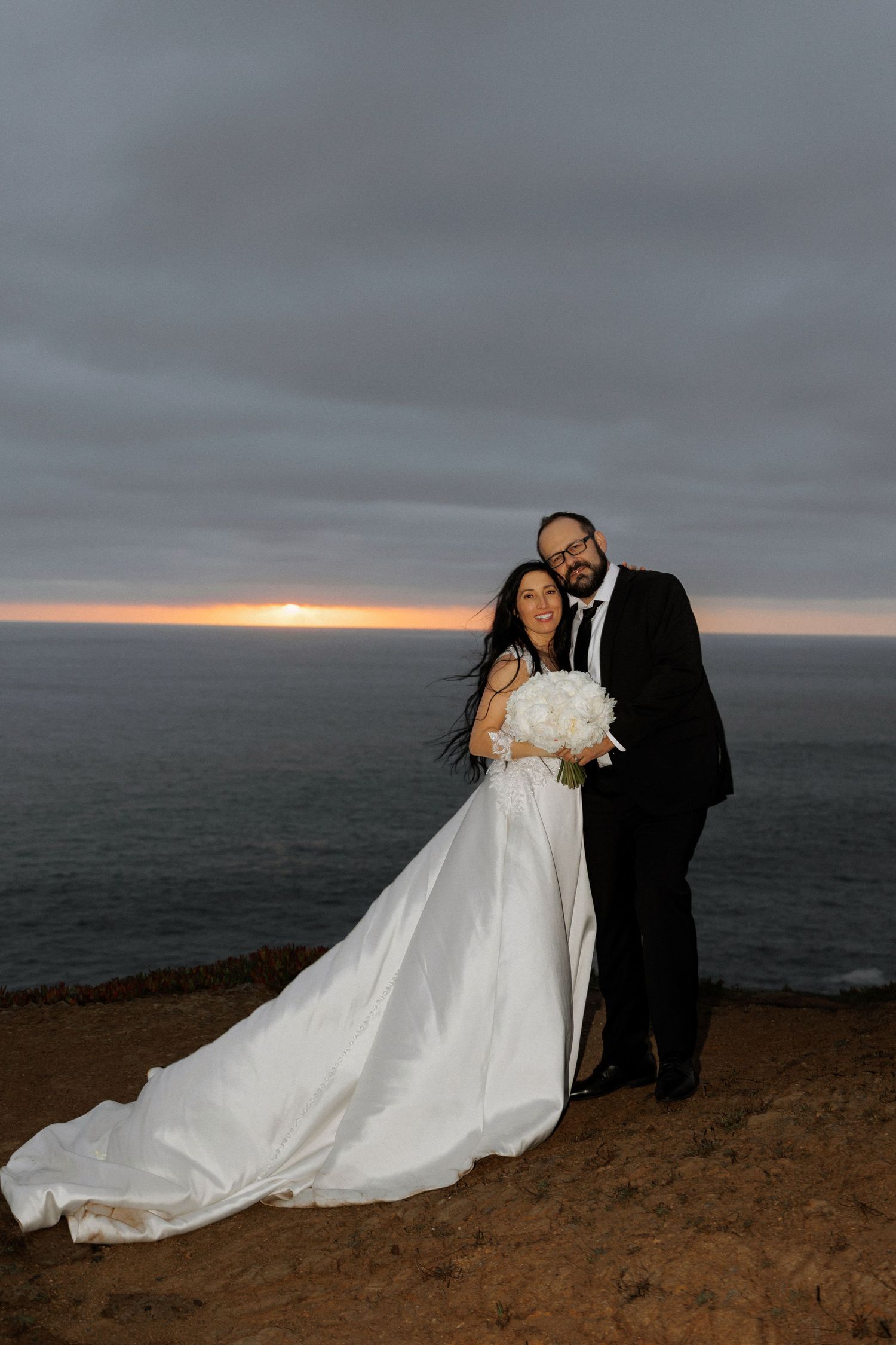 Wedding couple poses against dramatic coastal sunset with flowing white dress.