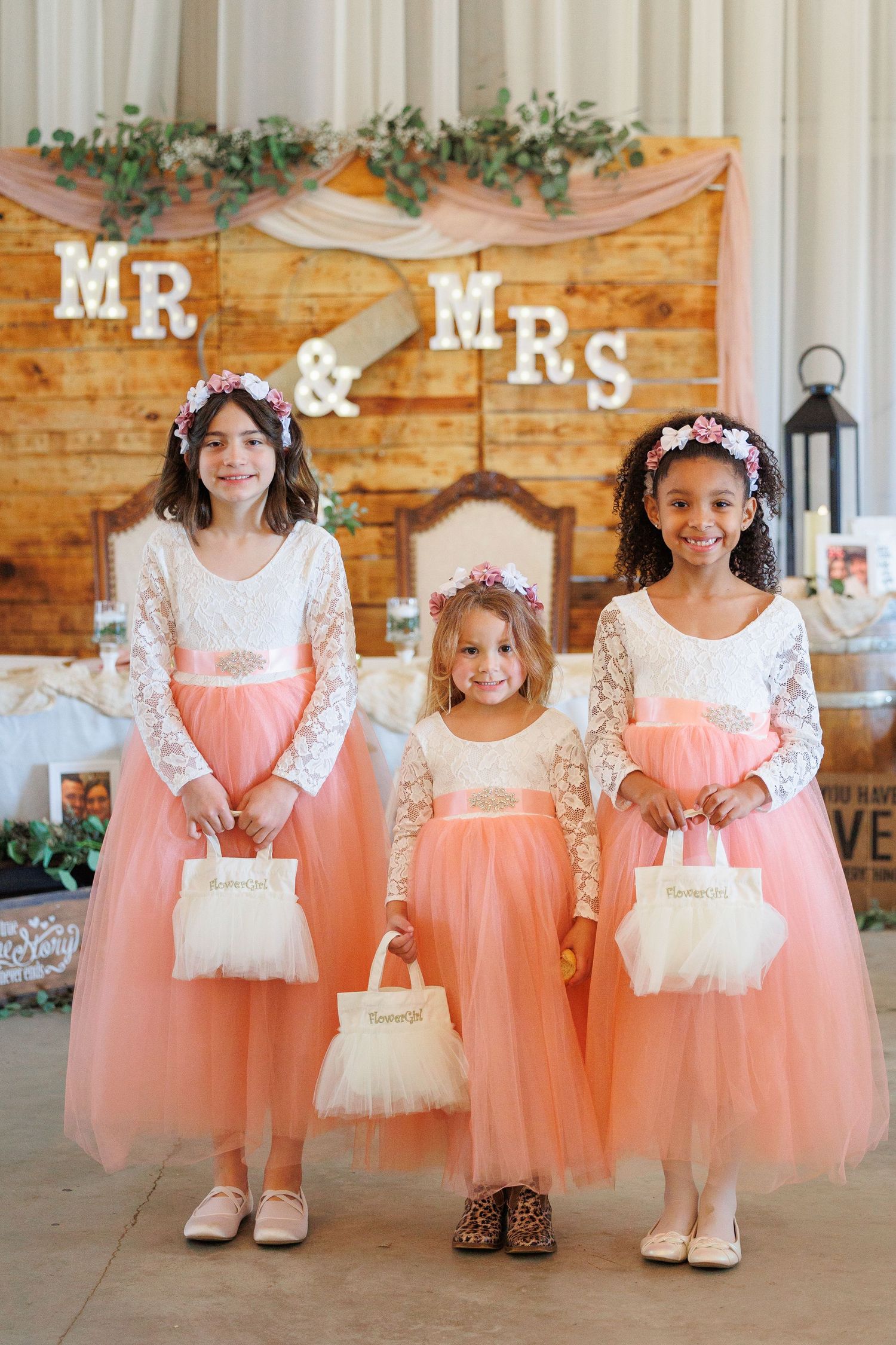 Young flower girls in peach tulle dresses and white baskets pose together at a rustic wedding venue.