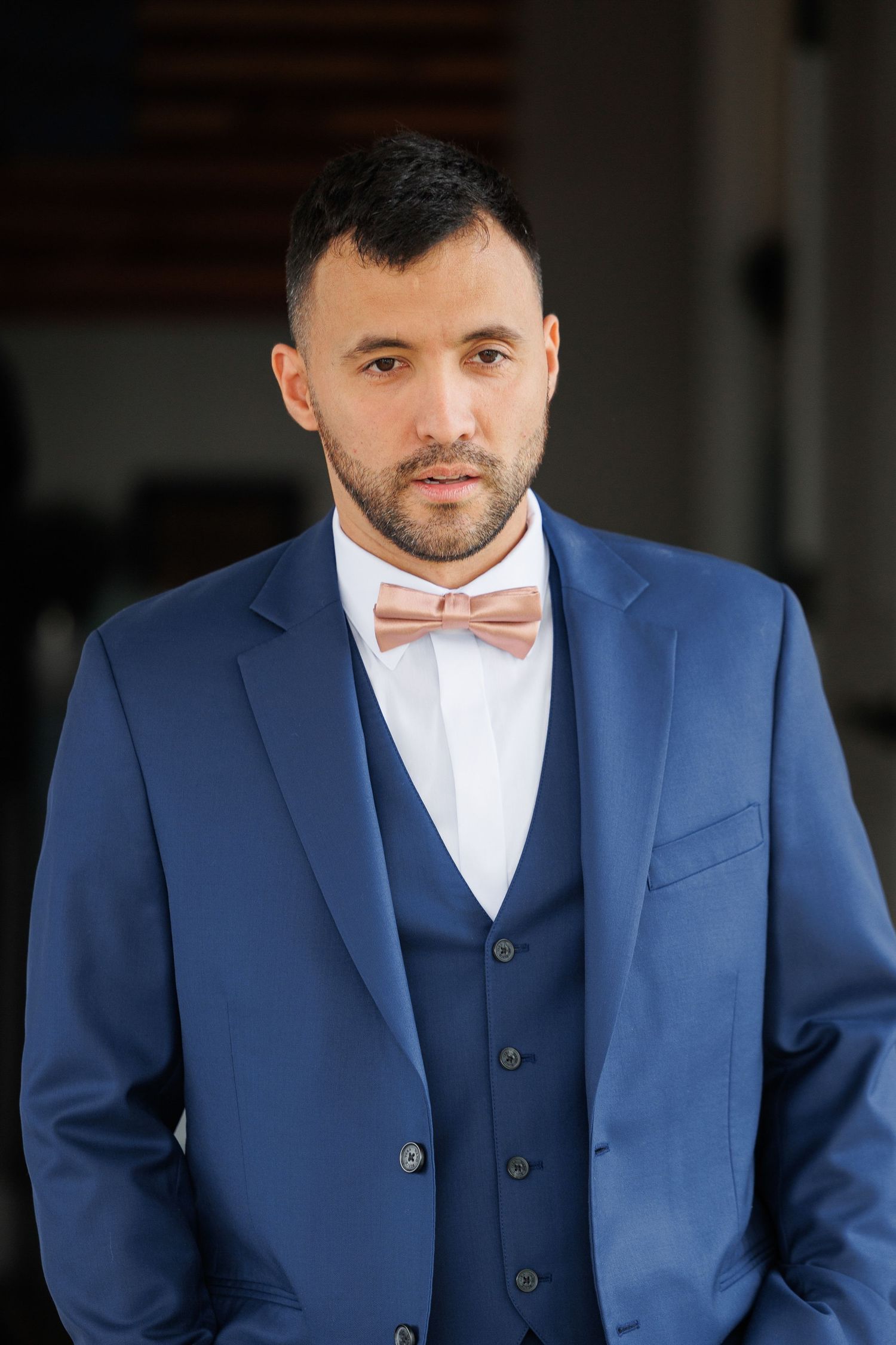 A person wearing a blue suit and pink bow tie poses in formal attire against a light background.