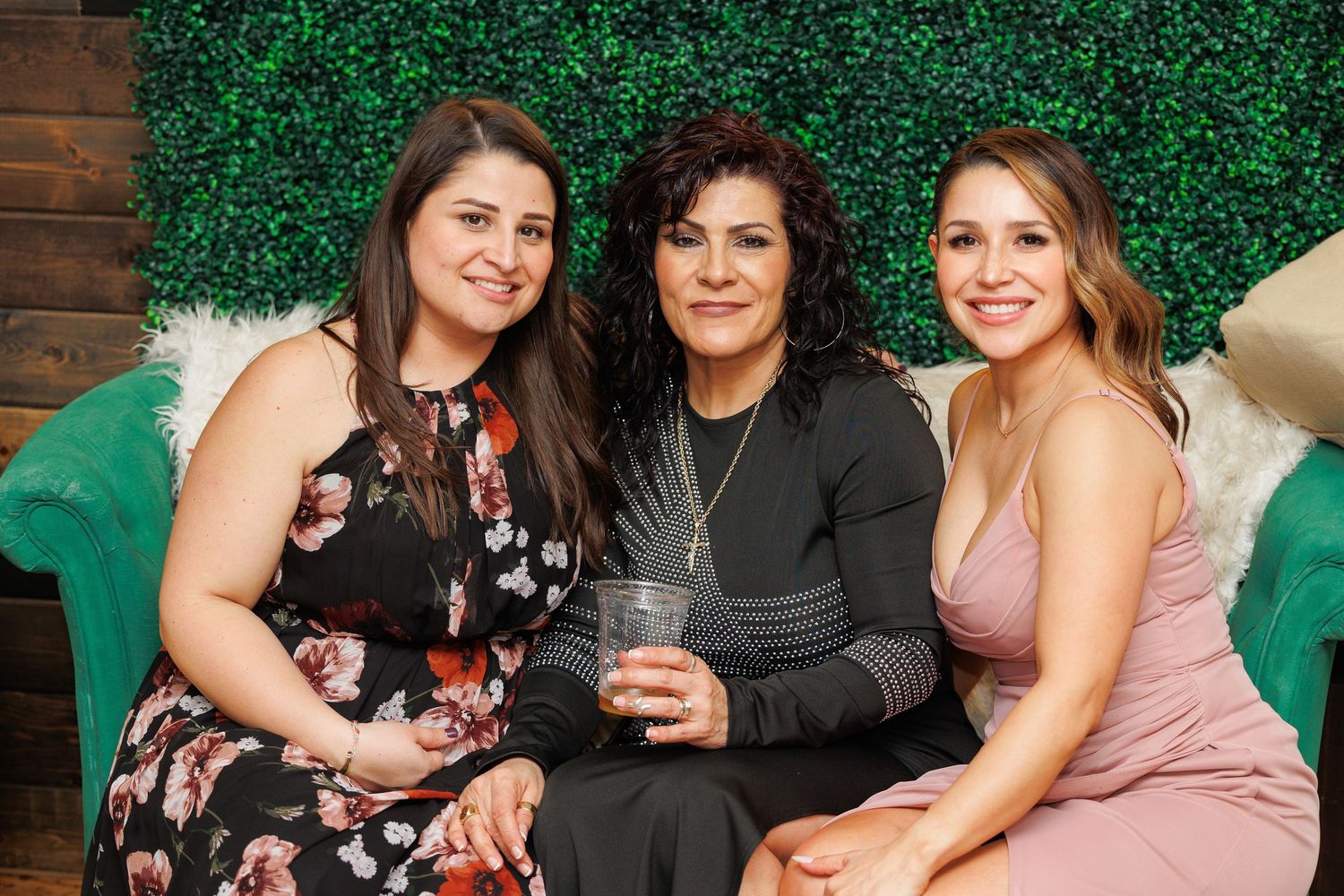 Three people pose together in front of a green hedge wall at what appears to be a social gathering.