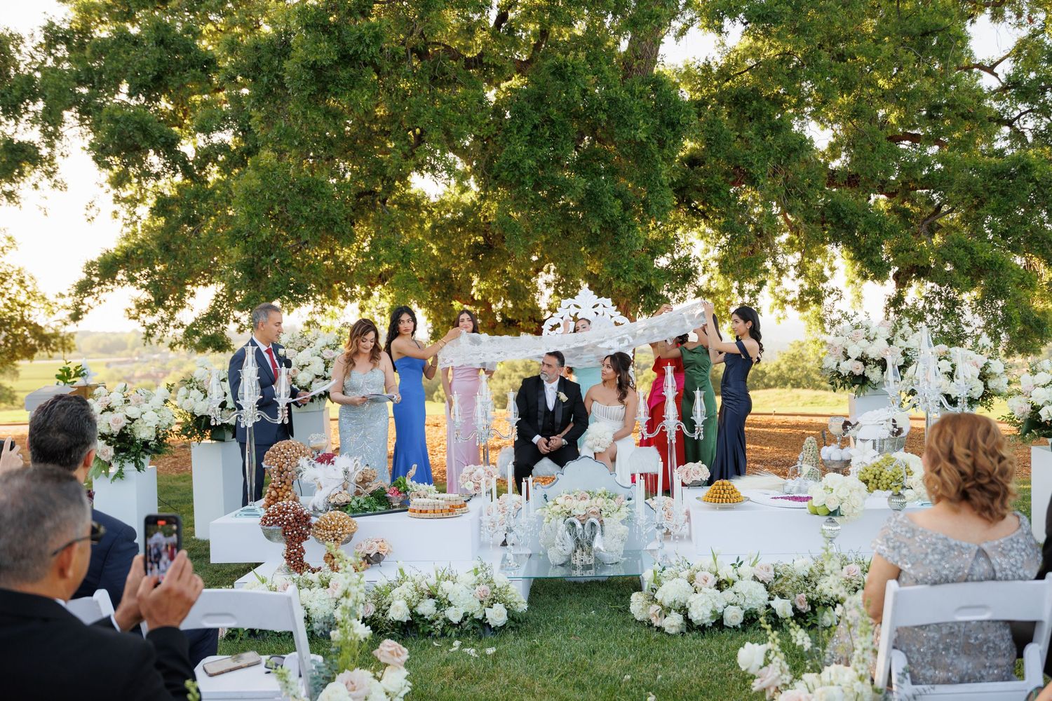 Elegant outdoor wedding reception setup with white chairs and tables arranged under green trees at sunset.