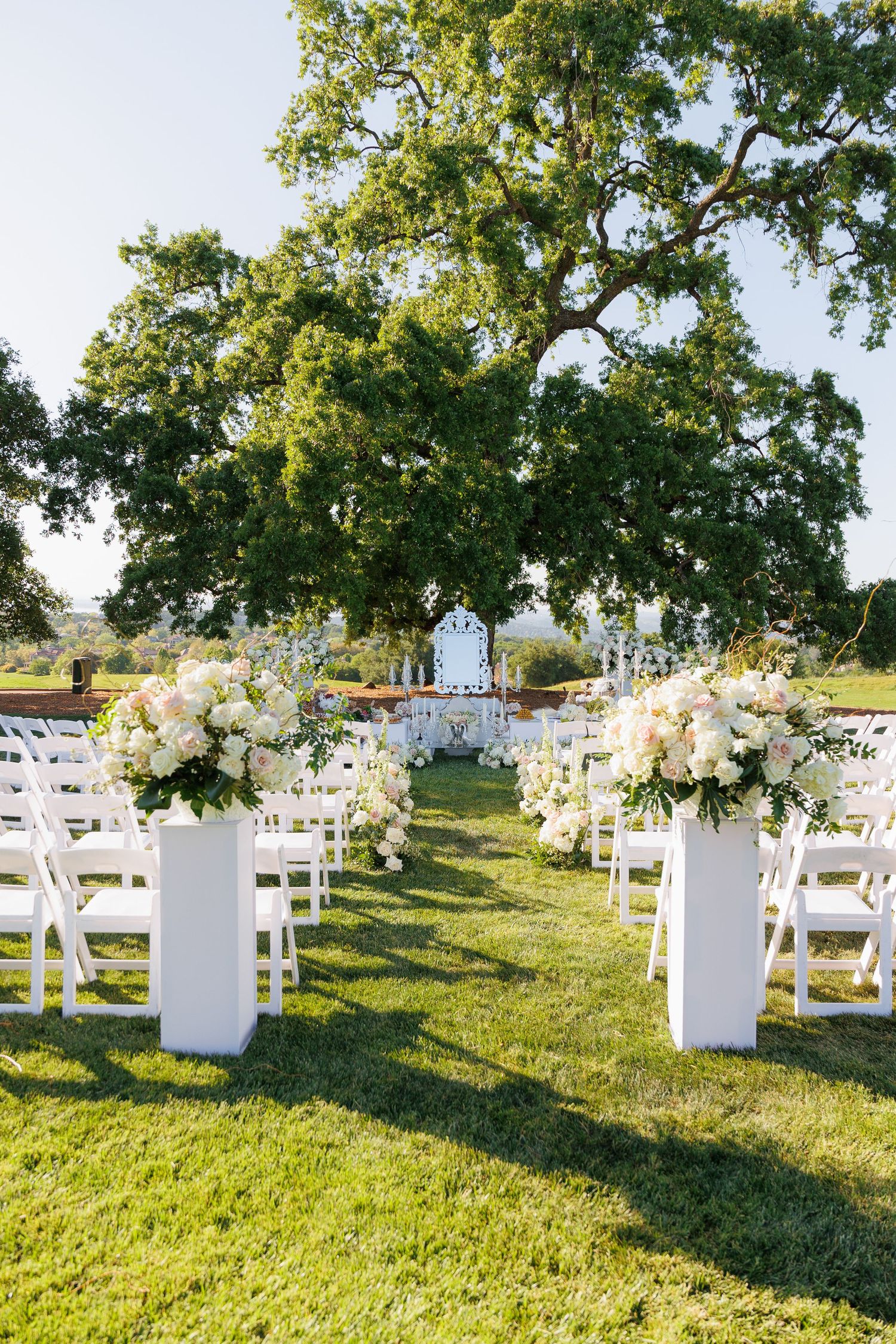 White wooden chairs arranged on a grassy hillside for an outdoor wedding ceremony under oak trees.