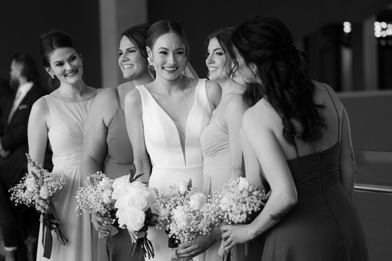 Bridesmaids in elegant light-colored dresses hold floral bouquets while sharing a candid moment at a wedding celebration.
