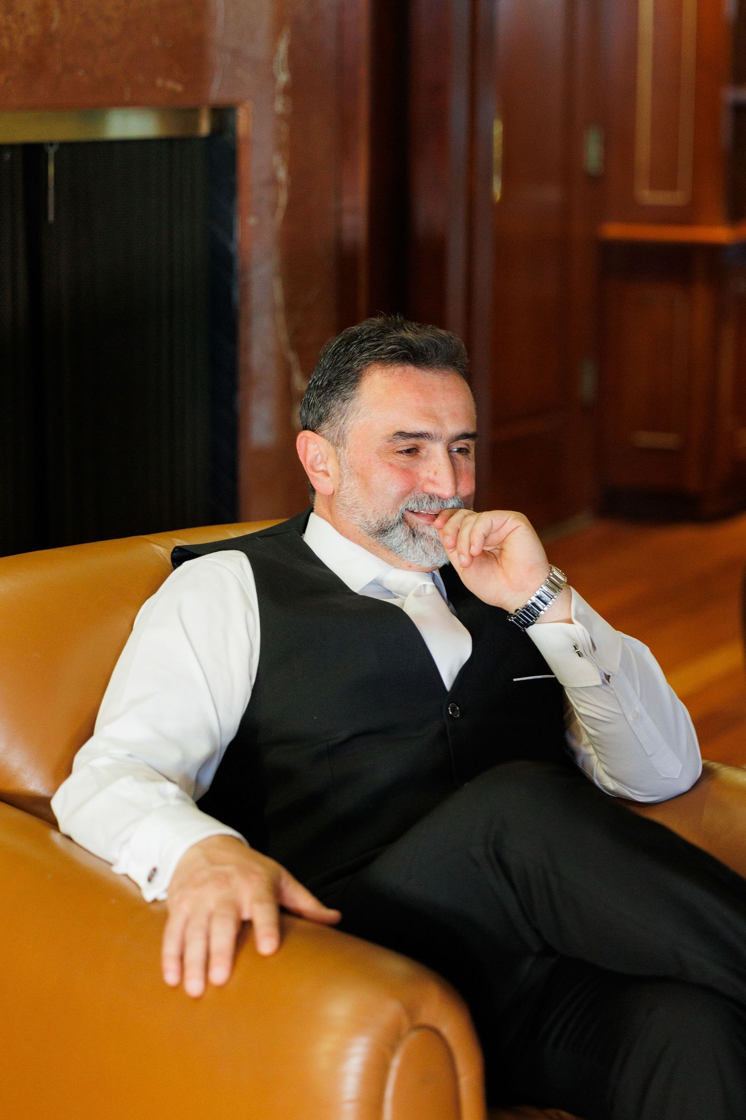 A person in a black vest and white shirt relaxes on a brown leather chair with a thoughtful expression.