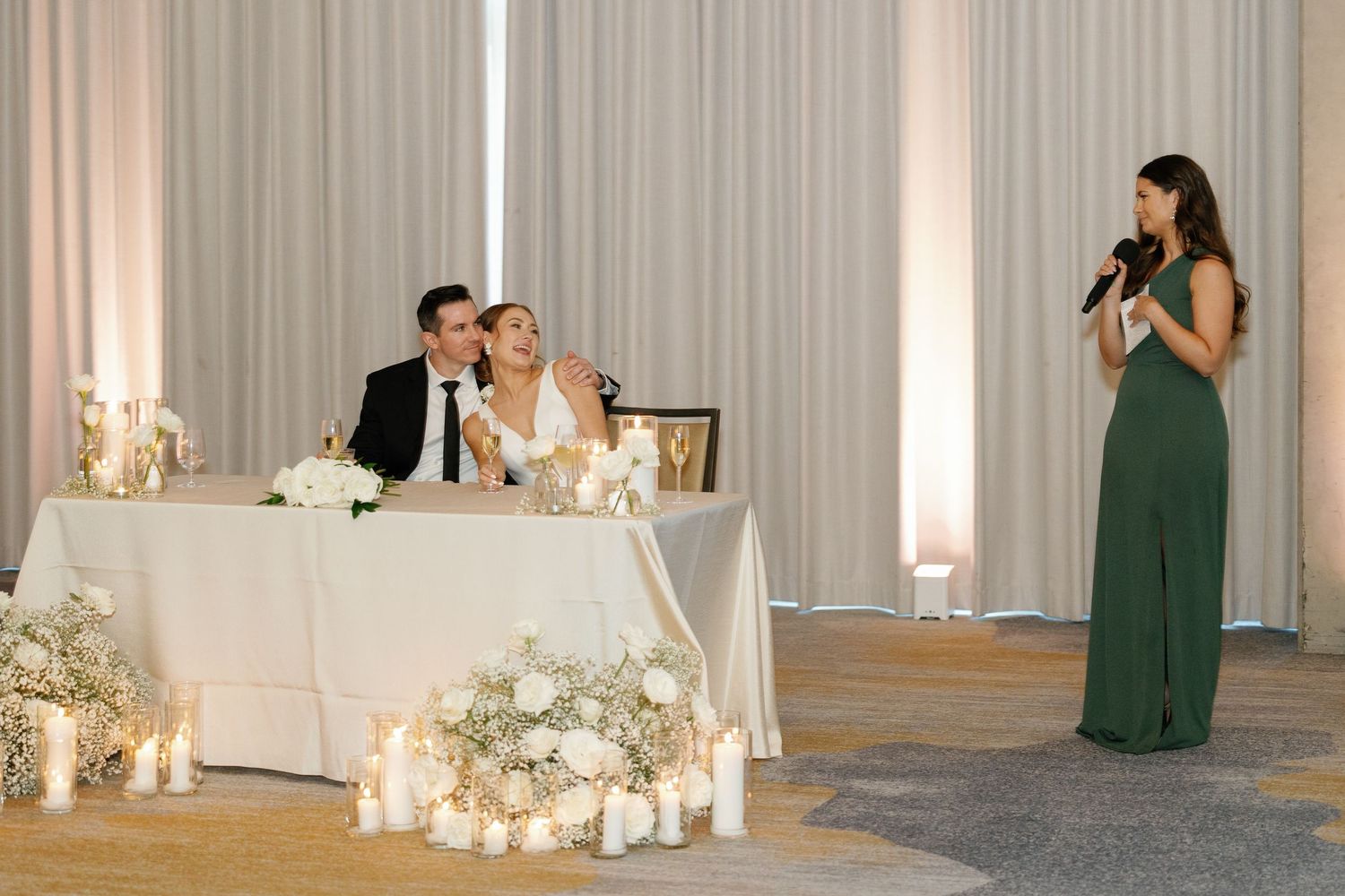 A wedding reception hall decorated with white curtains, candles and floral arrangements while guests mingle in emerald dresses.