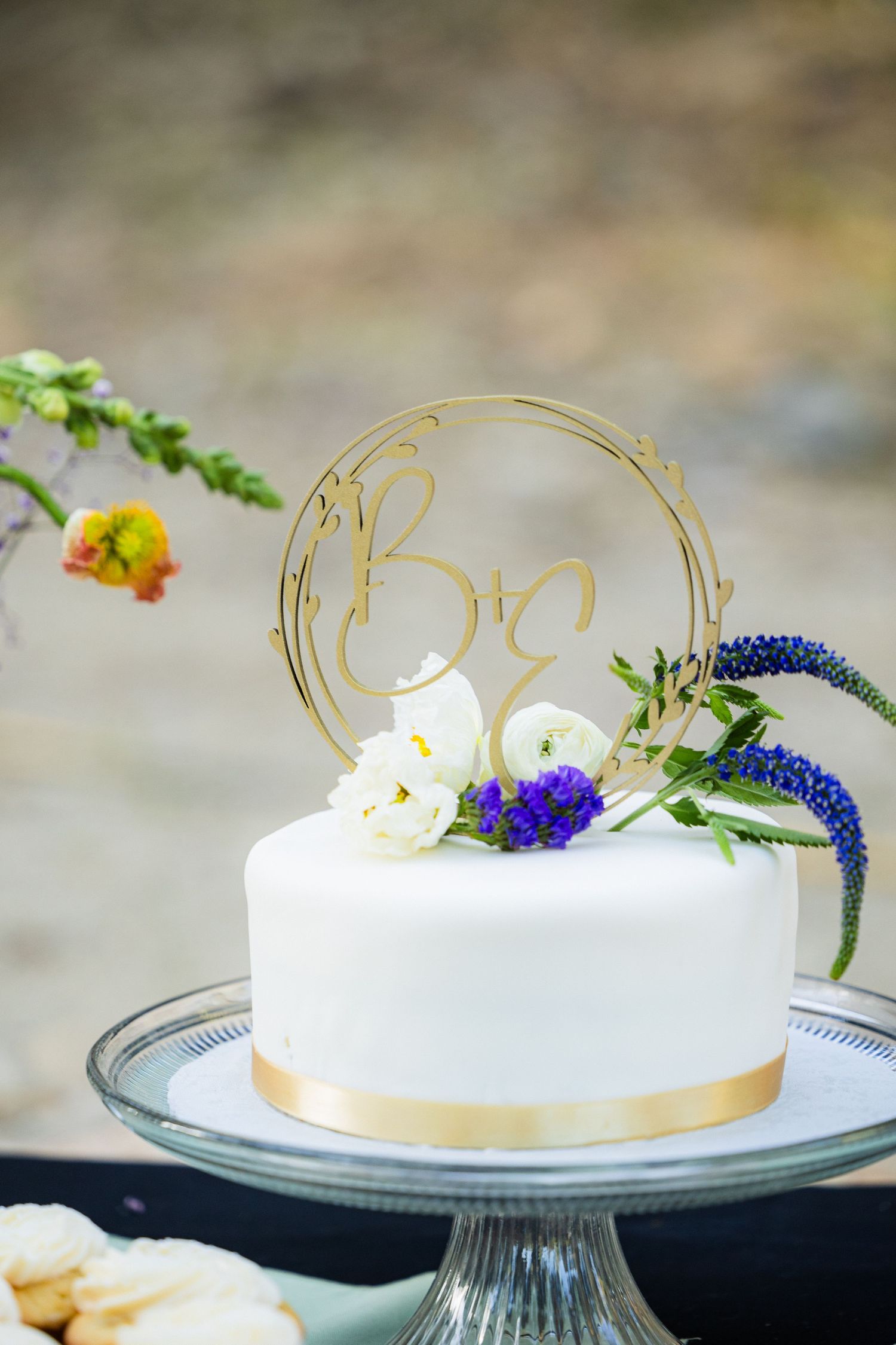 A white frosted cake decorated with blue flowers and gold accents sits elegantly on a glass cake stand.