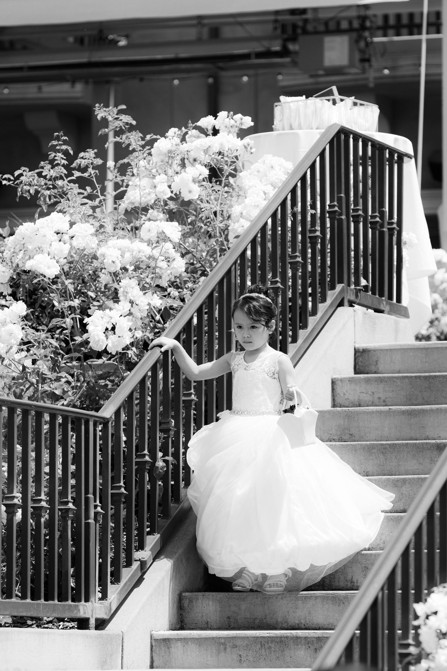 A young figure in a white flowing dress descends outdoor stairs adorned with blooming flowers in black and white.