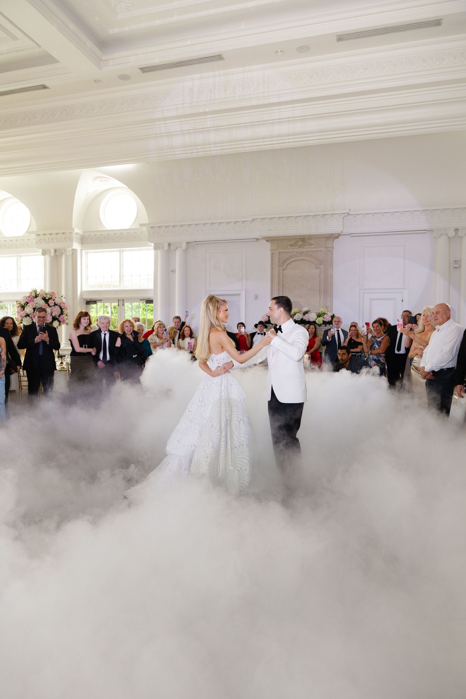 A couple shares their first dance on a misty dance floor with guests watching in an elegant white ballroom.
