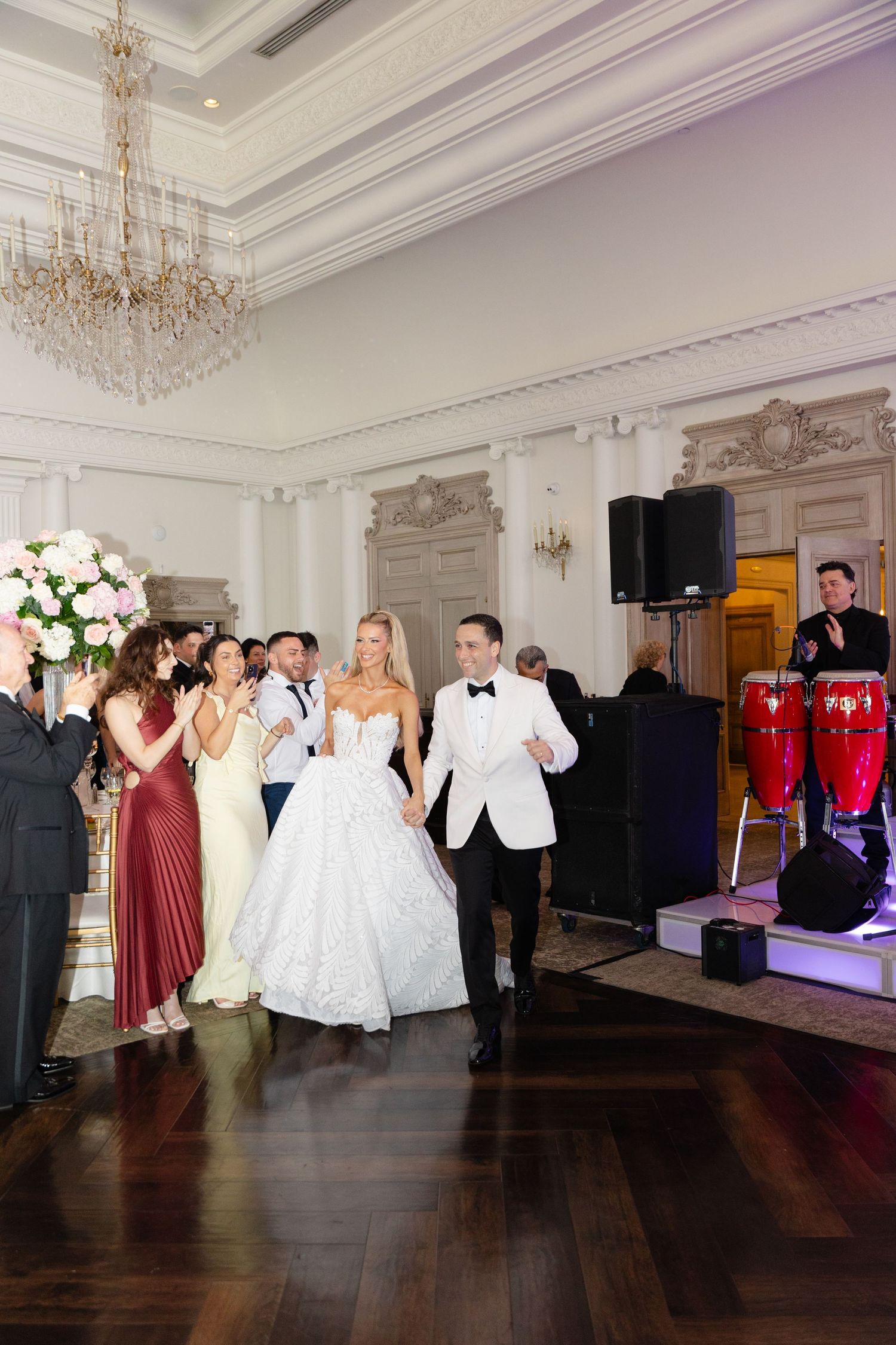 Elegant wedding reception in a grand ballroom with crystal chandelier, white decor and live band setup on stage.