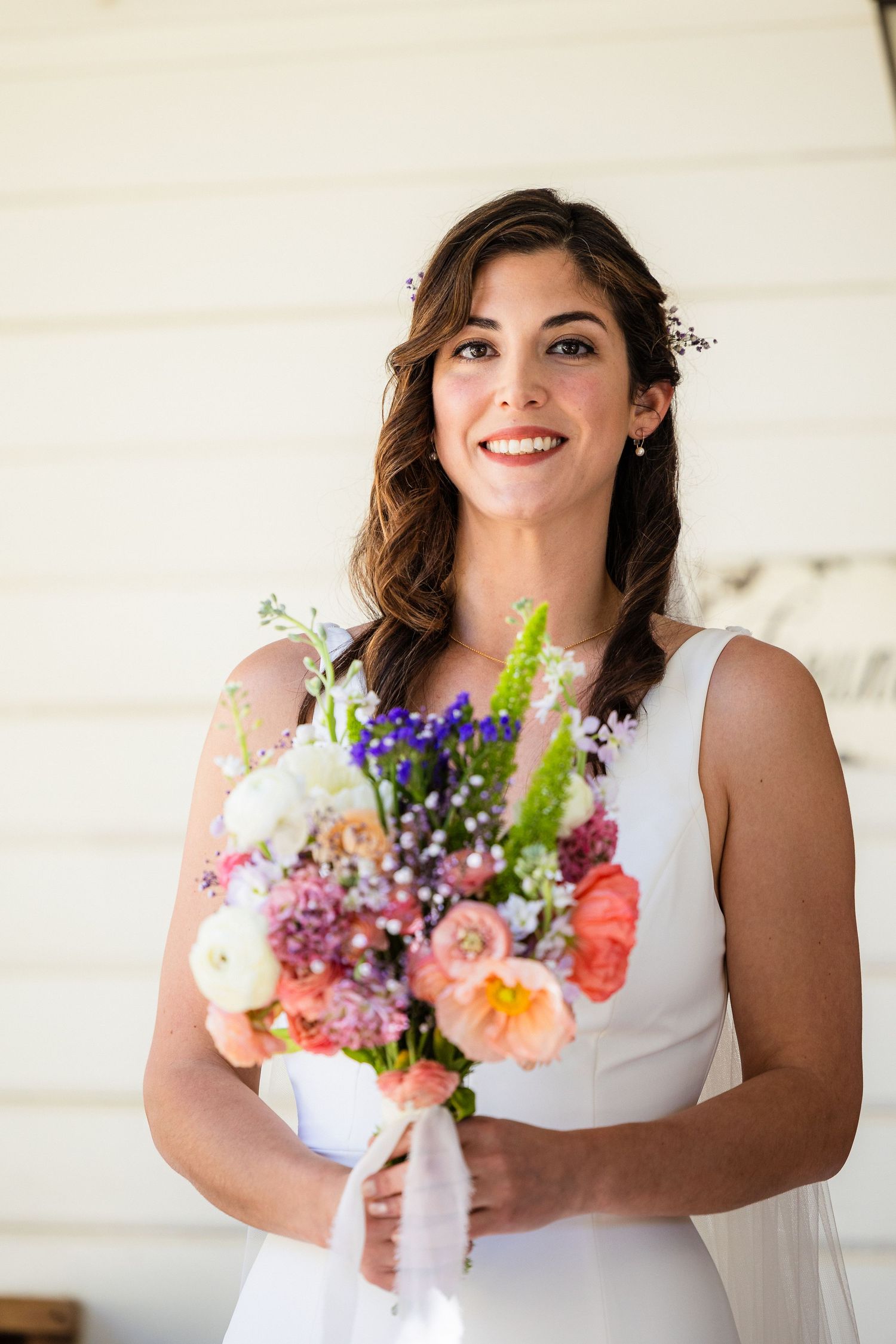 A bride in a sleeveless white dress holds a vibrant bouquet with coral, pink and purple flowers against a light background.