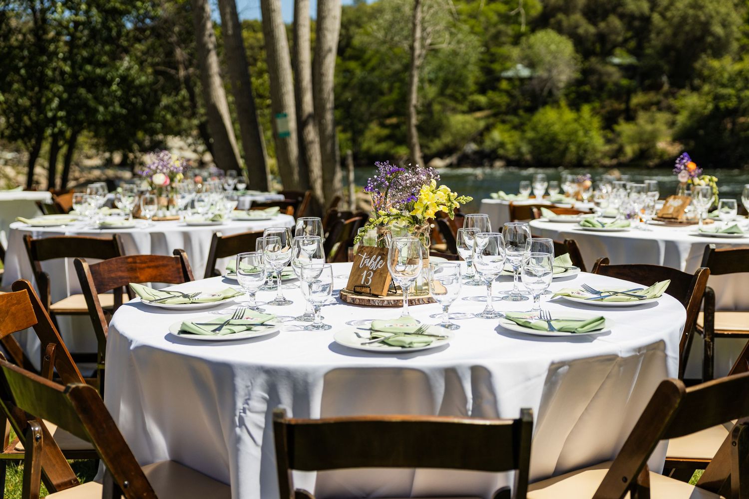 Elegant outdoor wedding reception tables with white linens and floral centerpieces set under tall trees in a garden setting.