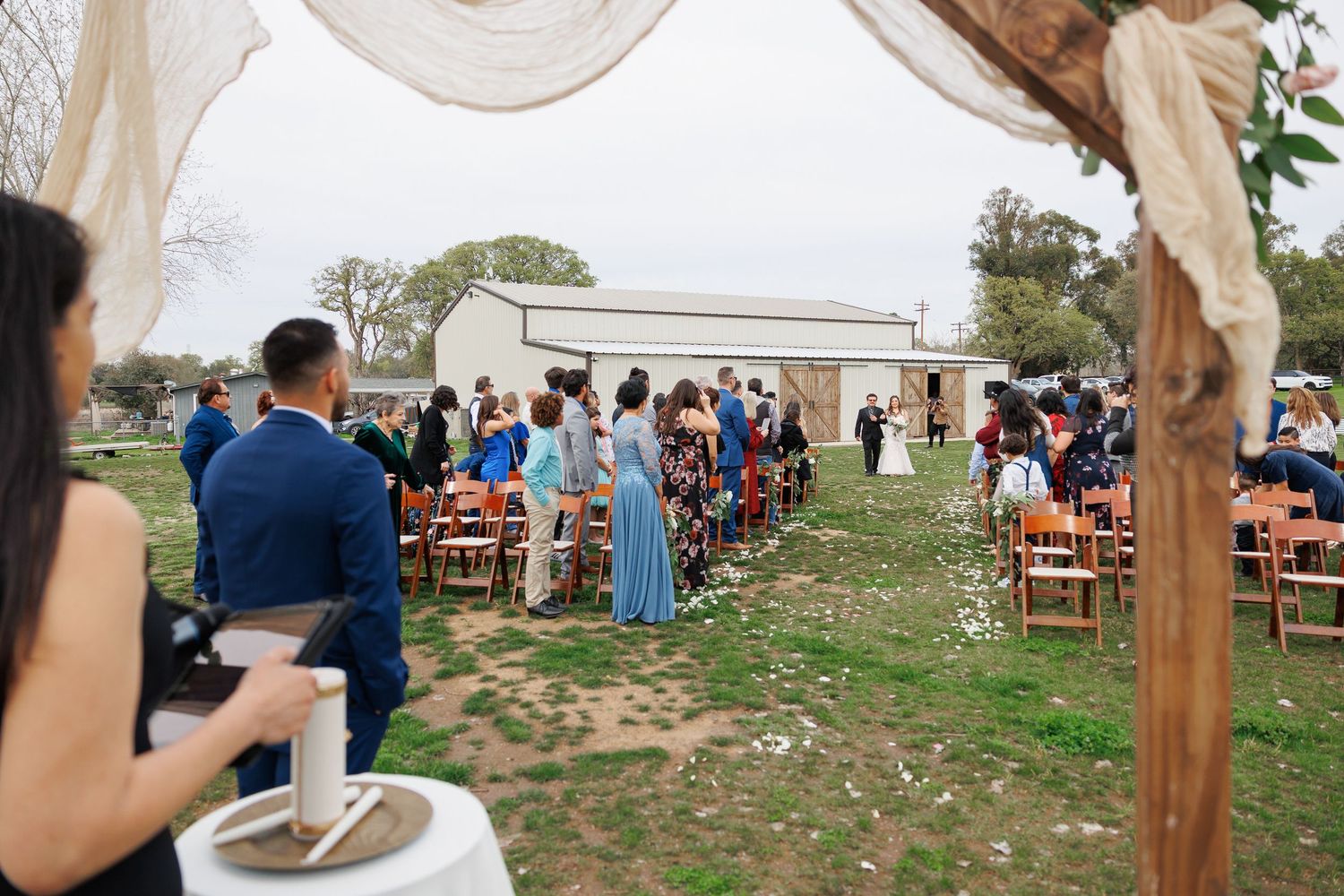 Outdoor wedding ceremony setup with wooden chairs arranged in rows beneath tall trees and a white tent canopy.