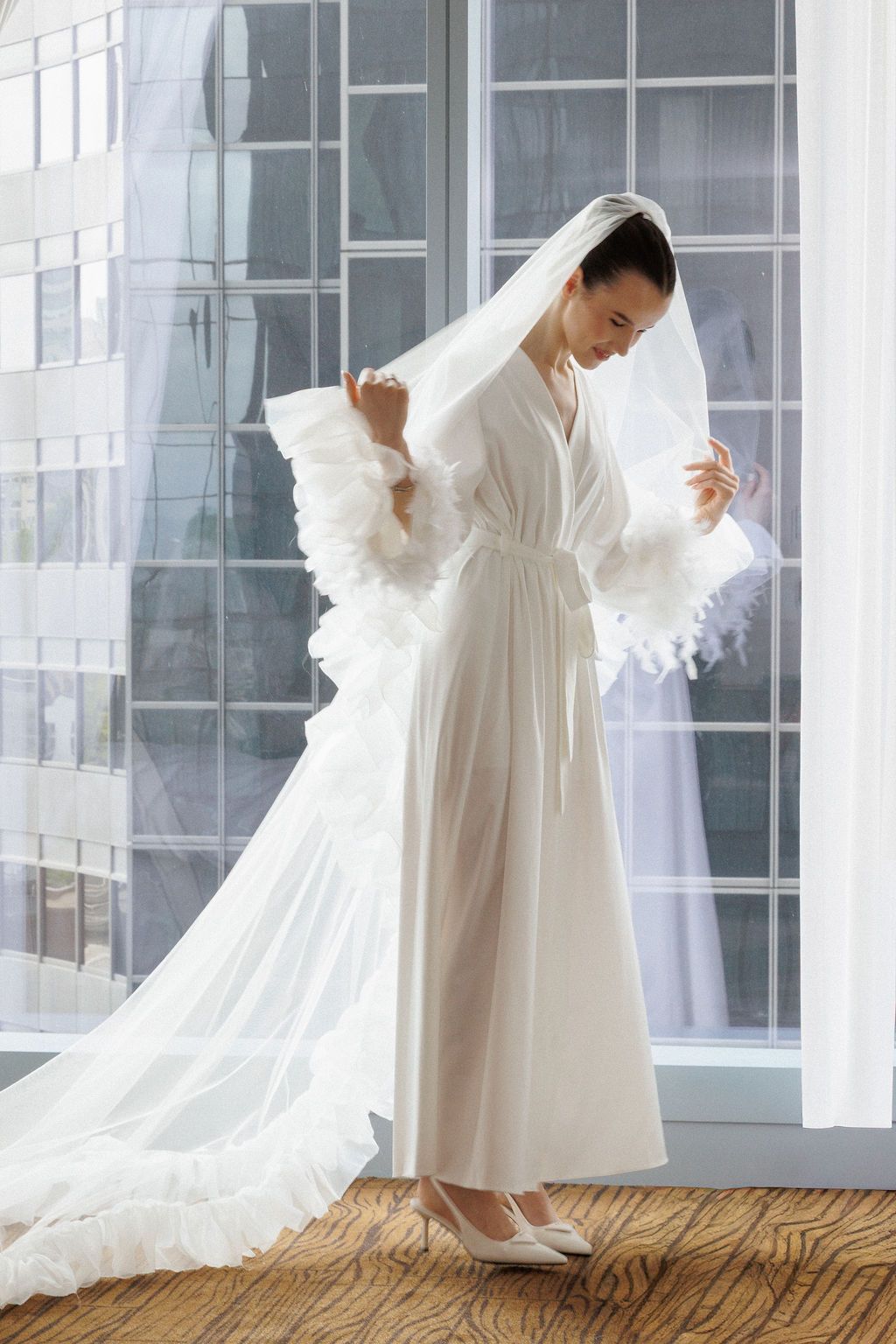 A figure in an elegant white robe and veil stands gracefully near windows in a bridal preparation setting.