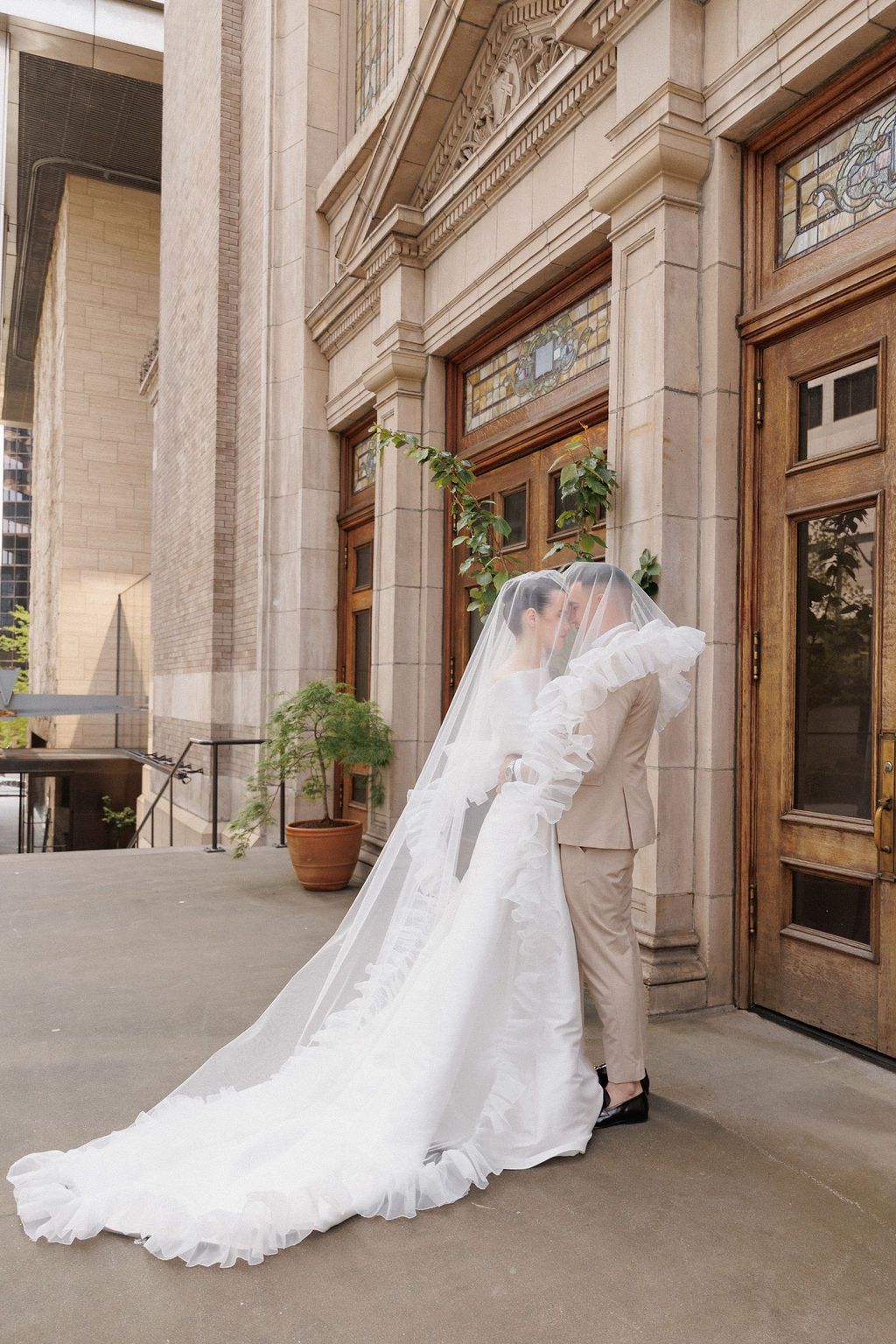 A dramatic wedding dress train and veil trails behind the couple as they walk through an ornate stone corridor.