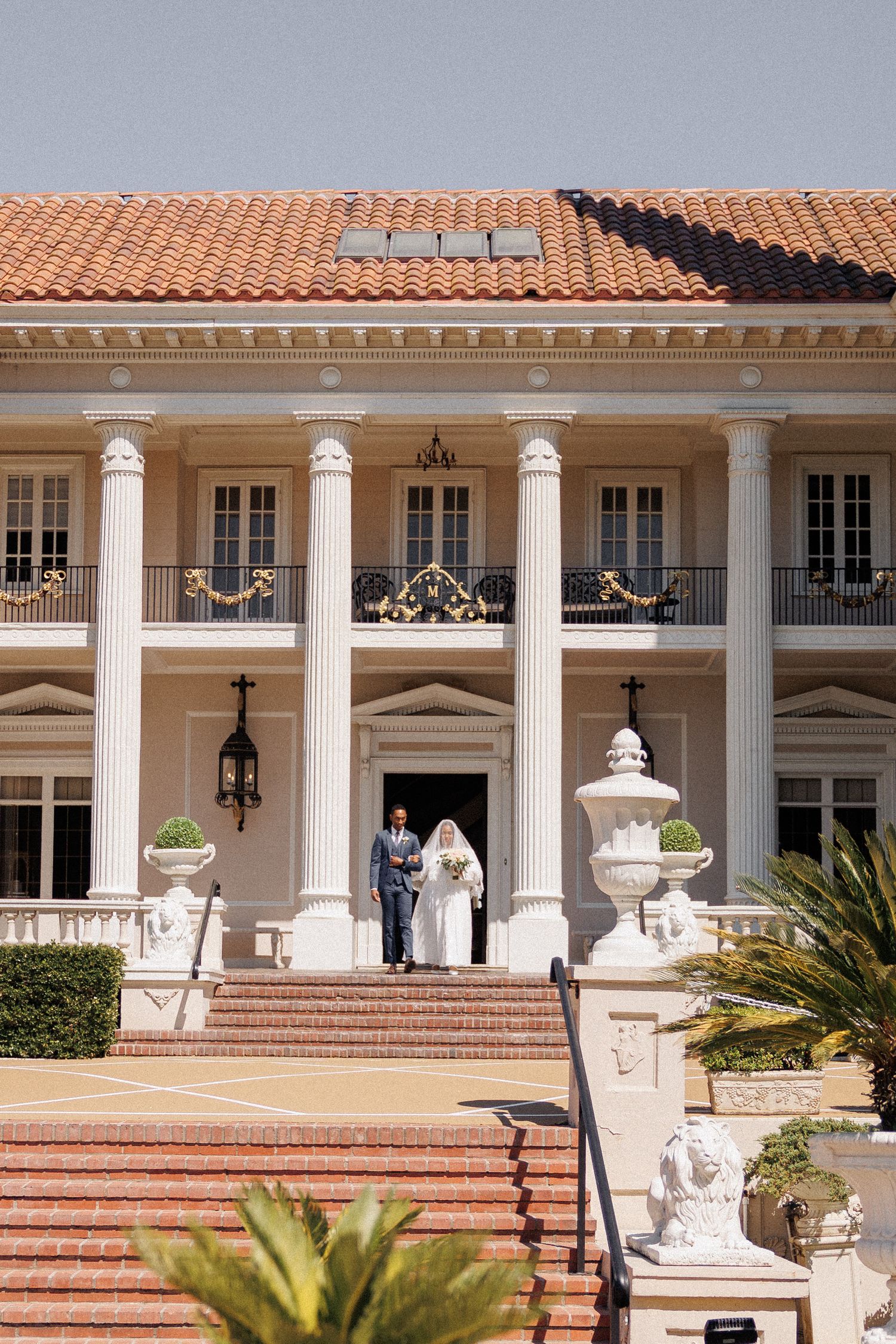 Elegant Mediterranean mansion entrance with white columns, terracotta roof, and grand staircase adorned with decorative urns.