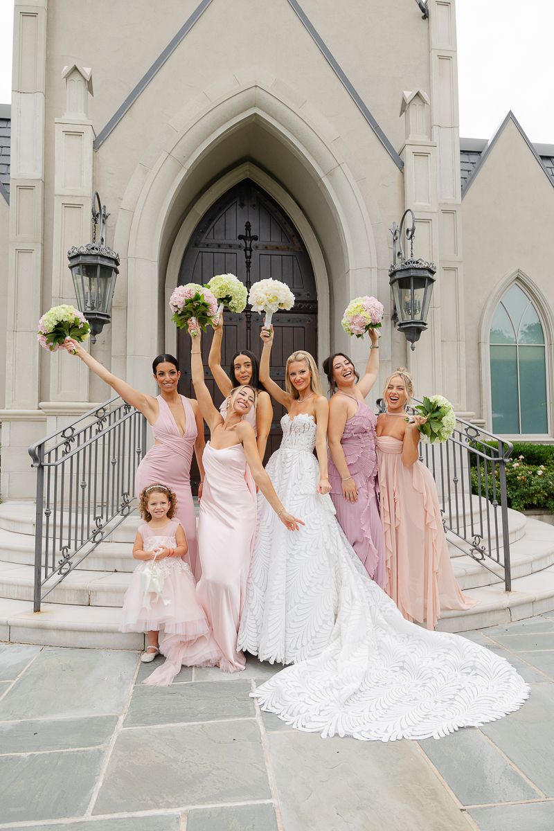 A bridal party poses joyfully on church steps with pink and purple dresses and white floral bouquets.