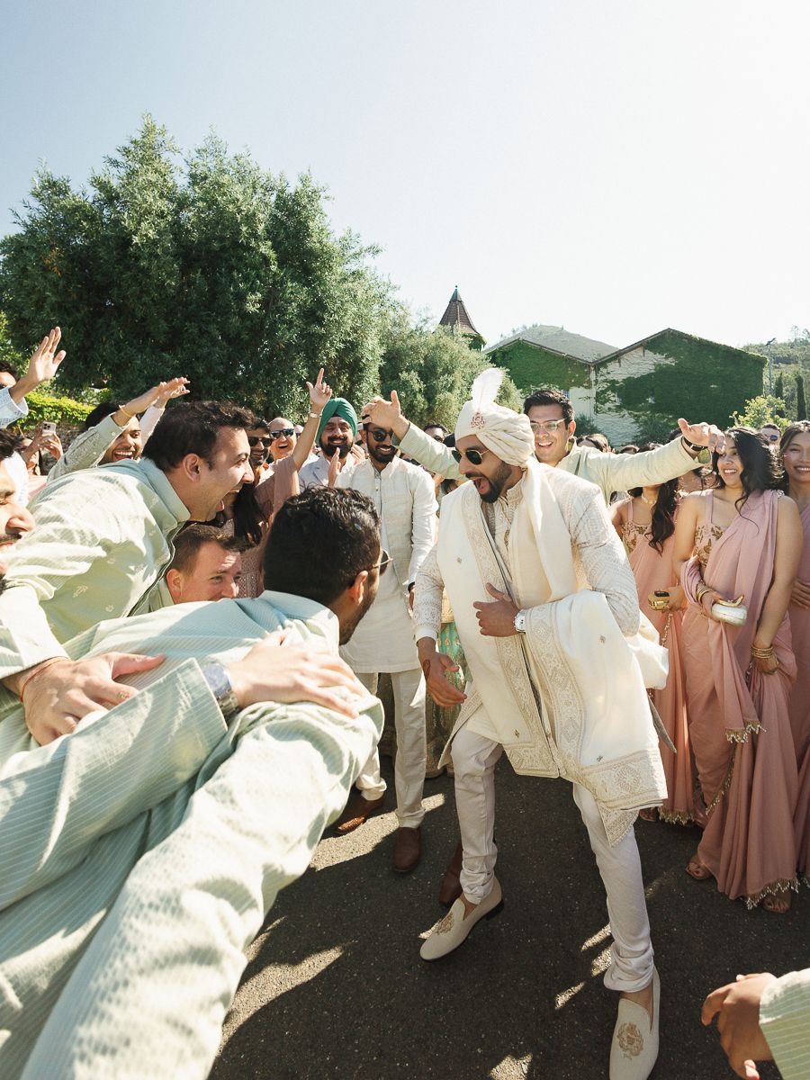 Wedding guests dance together in a circle wearing cream and blush colored outfits at an outdoor celebration.
