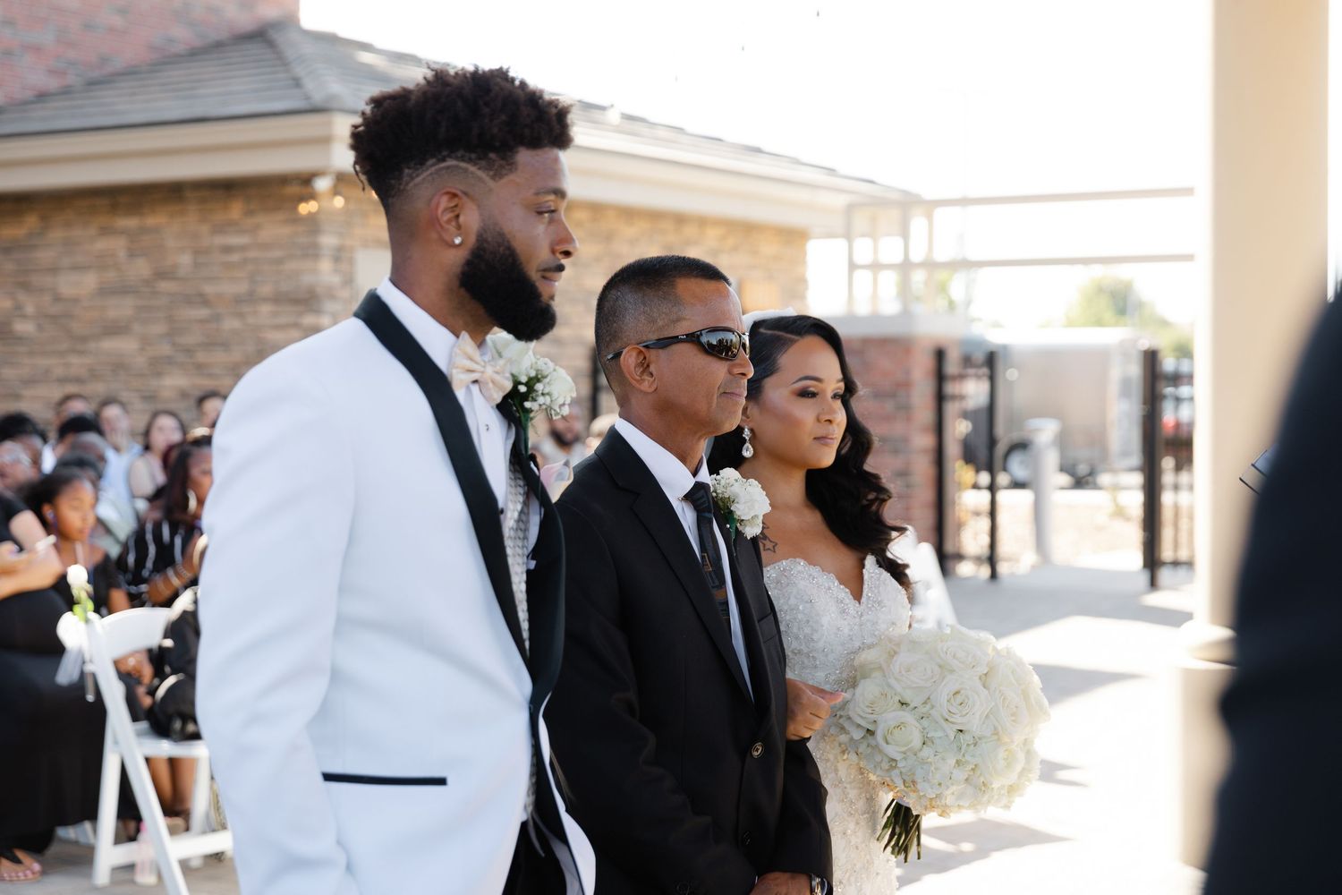 Wedding party members pose together during an outdoor ceremony with stone building and white chairs in background.