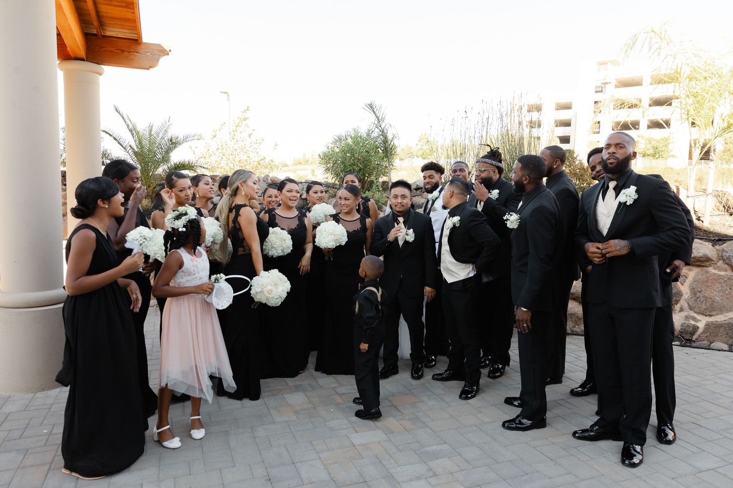 Wedding guests in formal black attire gather on an outdoor terrace during a formal celebration.