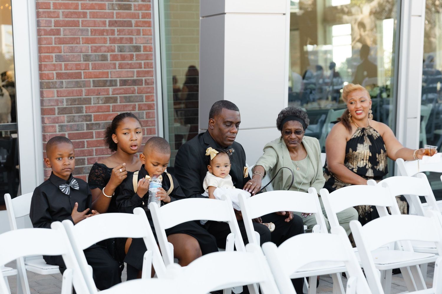 People seated in white folding chairs at an outdoor event venue with brick walls and windows in the background.