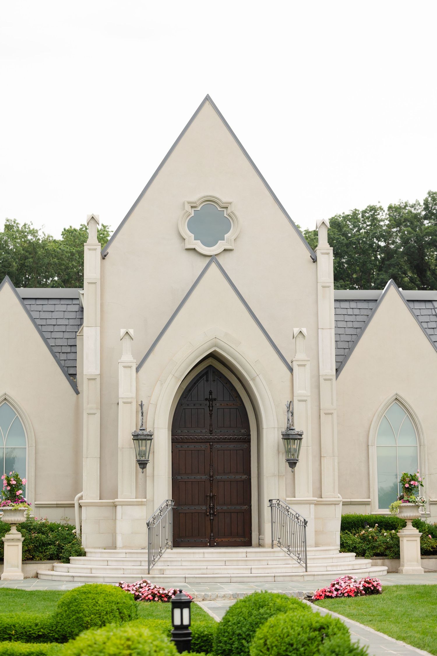 Three identical Gothic-style church entrances with arched wooden doors, white stone facades, and pink flowers in landscaping.