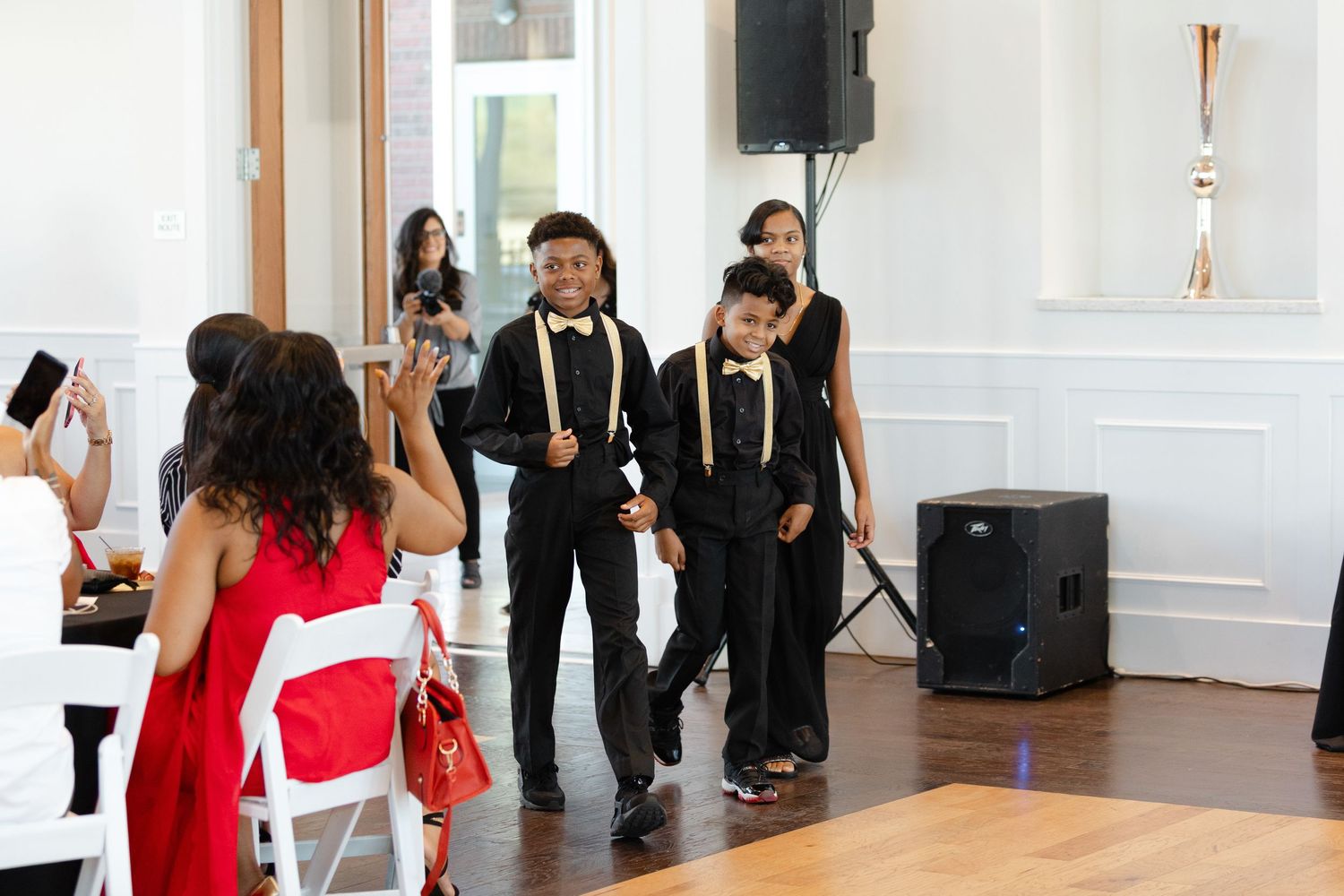 Wedding party members in formal black suits and bright red dresses pose together in an elegant white room.