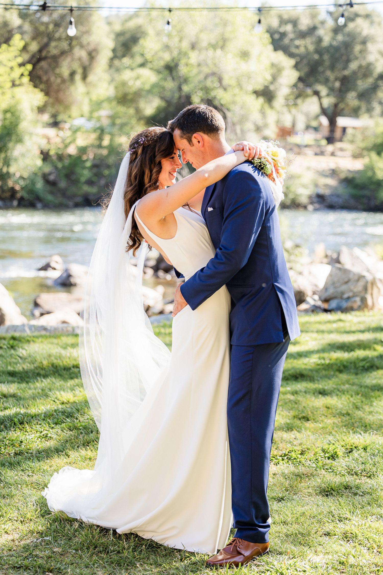 A couple shares a romantic dance together outdoors on grassy lawn by a lake during their wedding celebration.