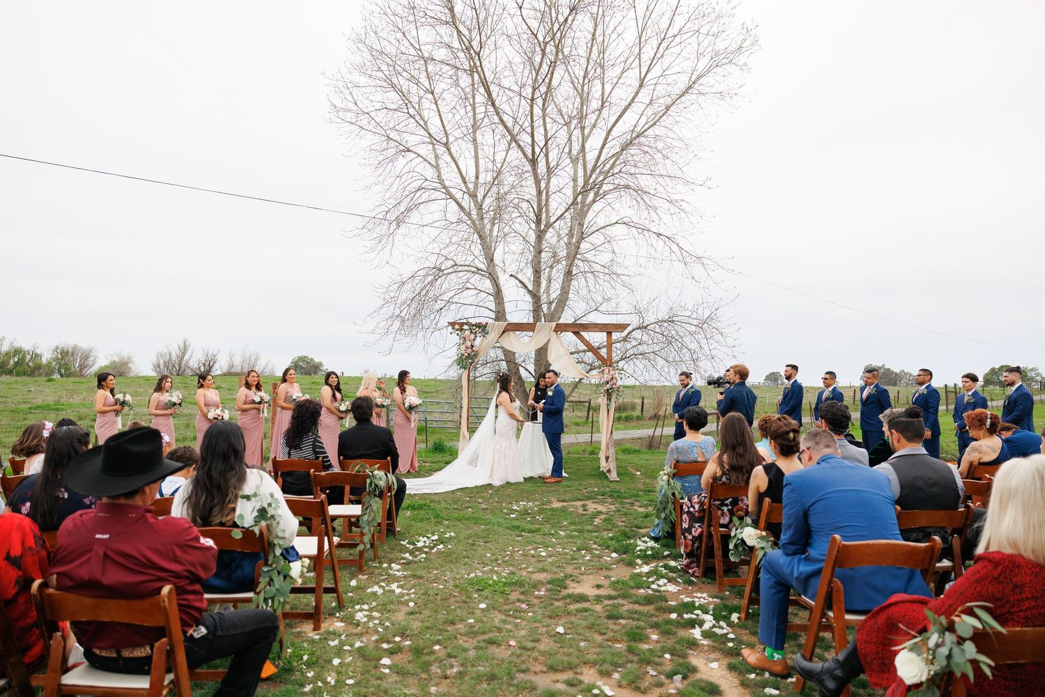 Rustic outdoor wedding ceremony with wooden chairs arranged on grass beneath a bare tree and wooden arch.