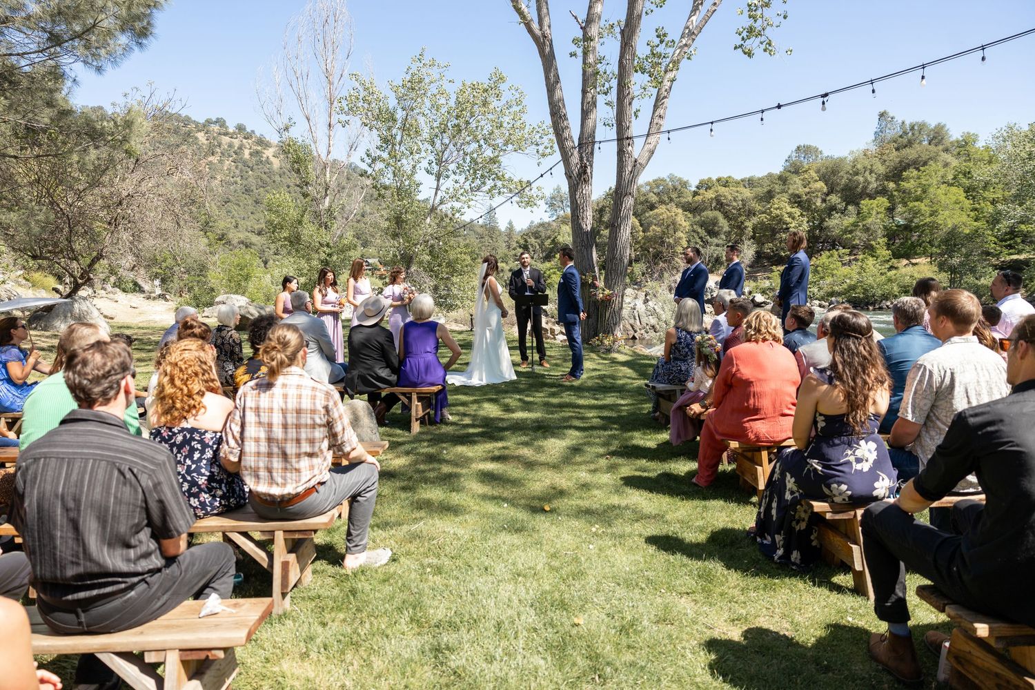 People enjoying an outdoor gathering with string lights and picnic tables in a scenic natural setting during daytime.