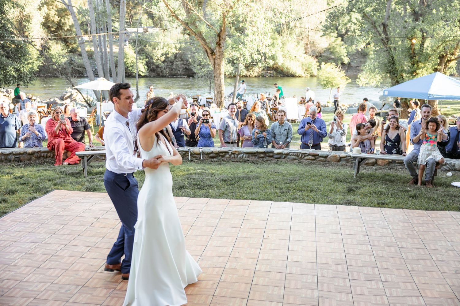 A couple in wedding attire dances together outdoors during their first dance at a summer wedding reception.