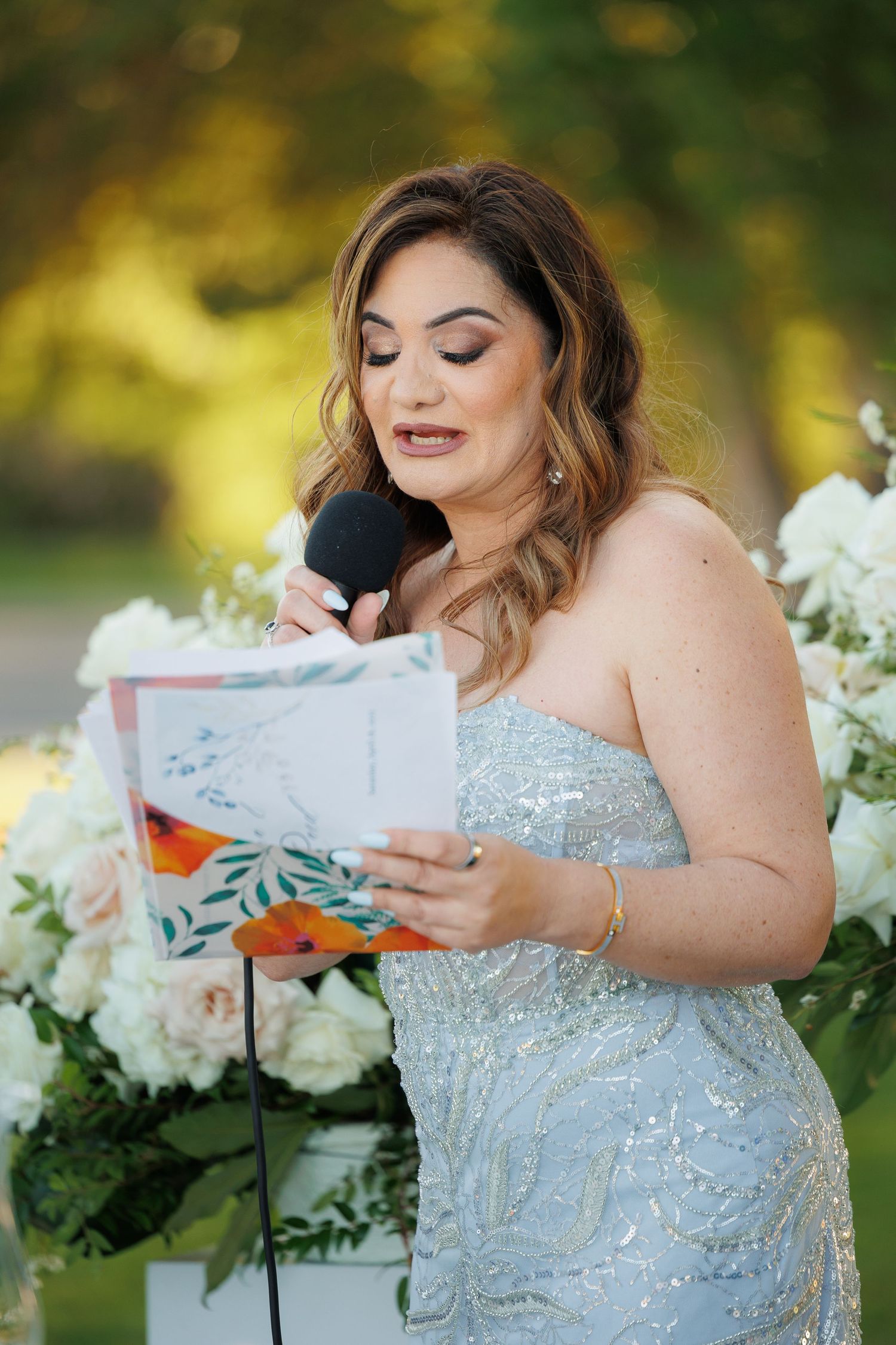 A bride in a sparkly strapless gown reads a letter outdoors with white floral arrangements in the background.