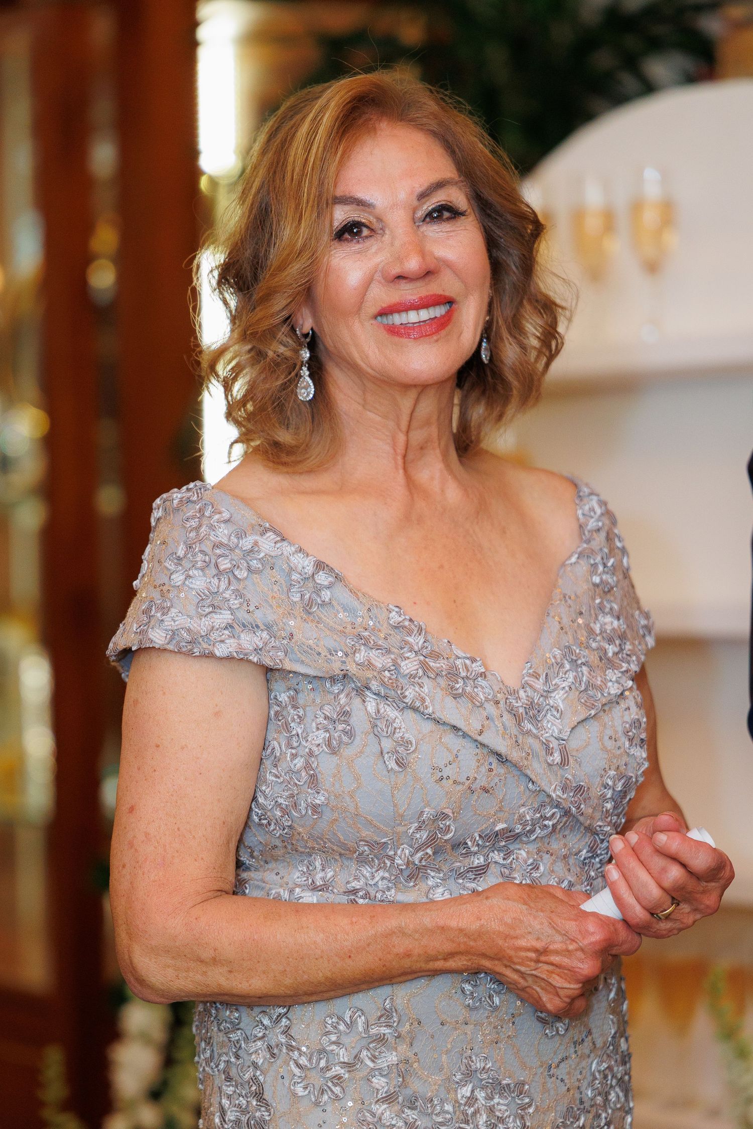 Woman in elegant silver beaded evening gown with cap sleeves and v-neckline smiles warmly at formal event.