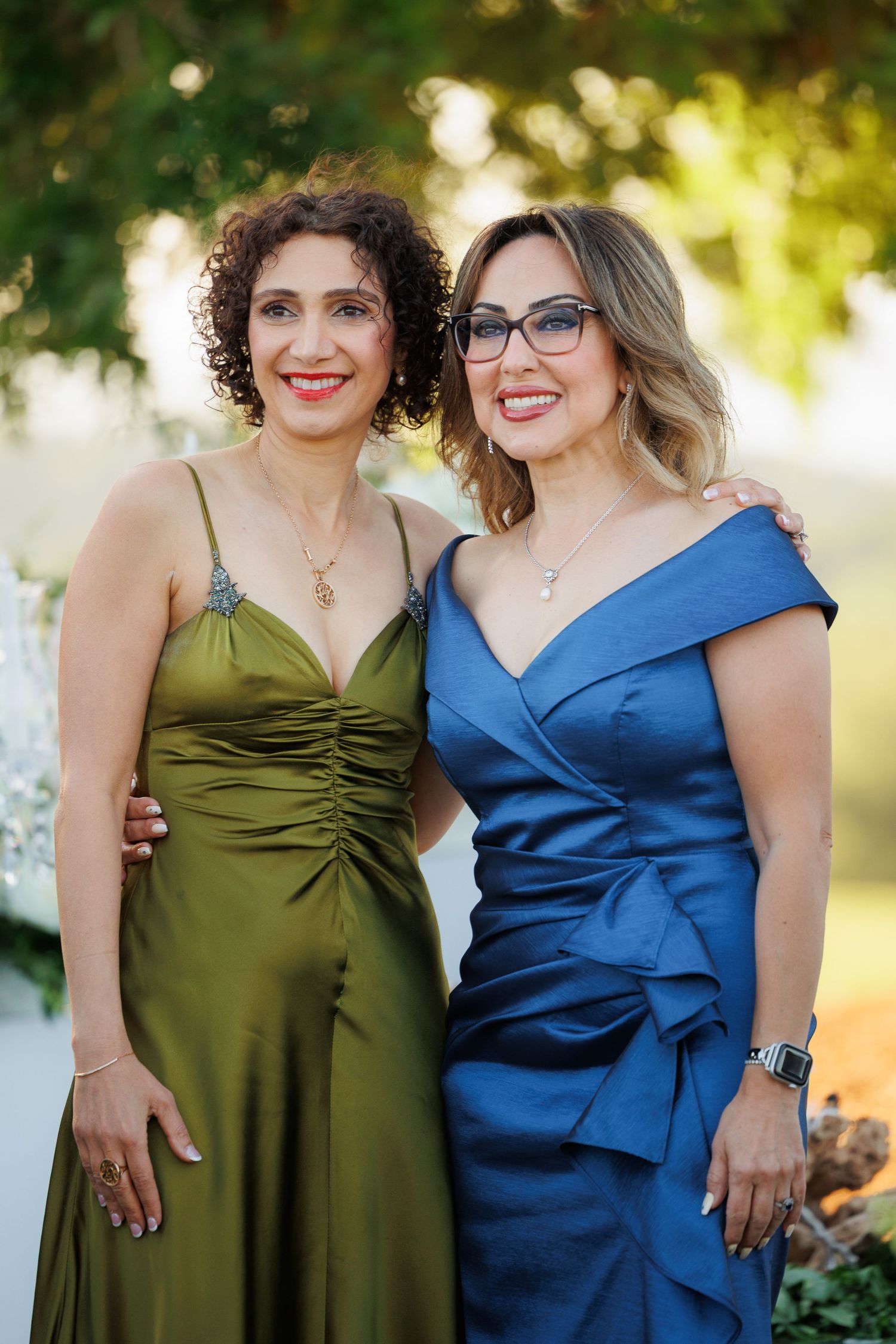 Two people in elegant dresses, one wearing olive green and one in royal blue, pose together outdoors at sunset.