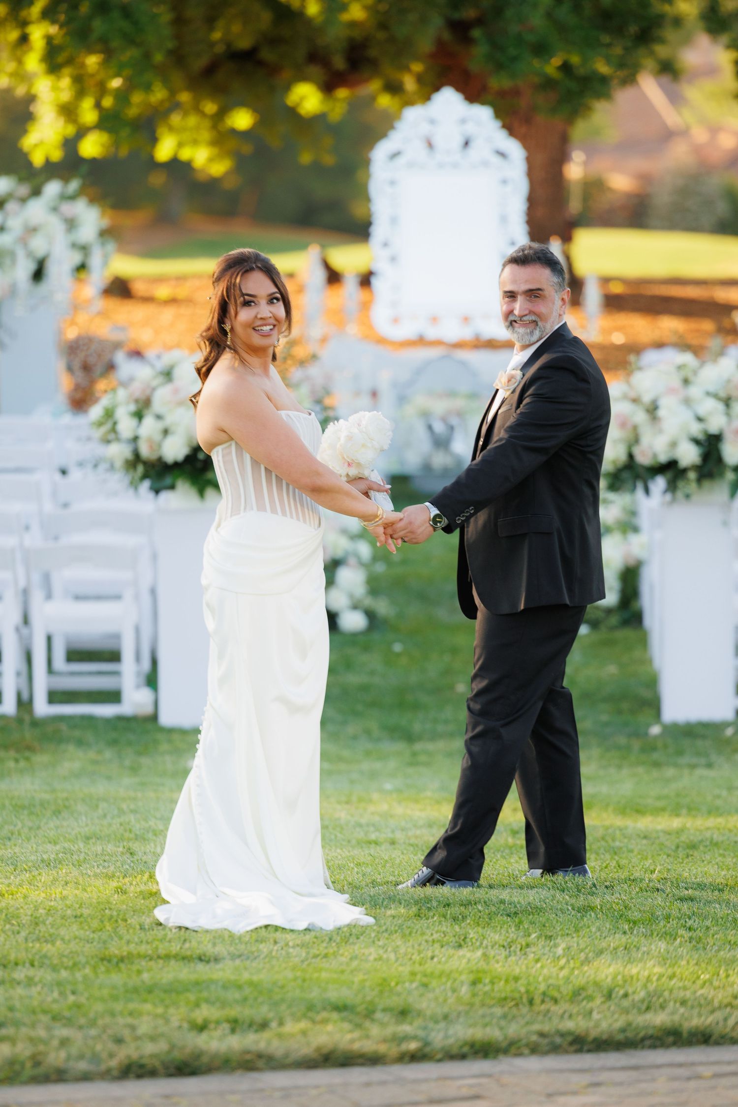 A couple in wedding attire playfully dance and twirl on a lawn with white chairs and floral arrangements in the background.