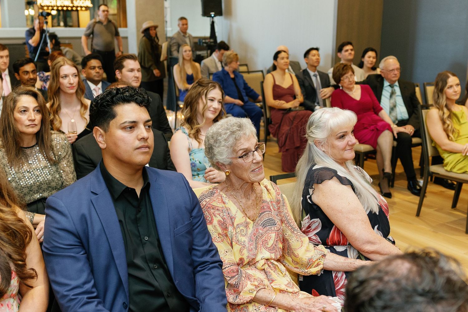 Wedding guests seated in rows at an indoor ceremony venue with blue walls and wooden floors.