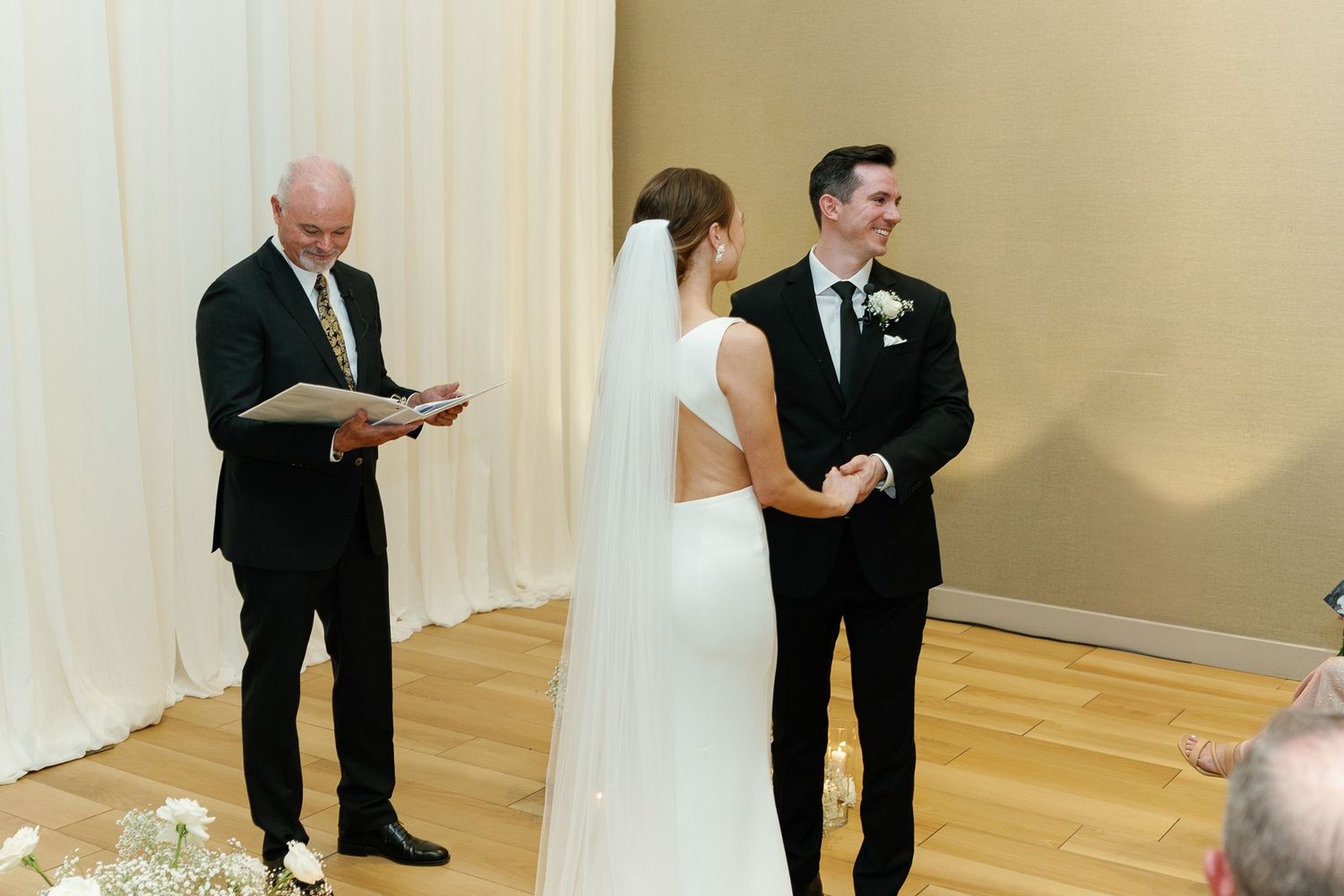 A wedding ceremony takes place in a simple indoor venue with light colored walls and hardwood floors.