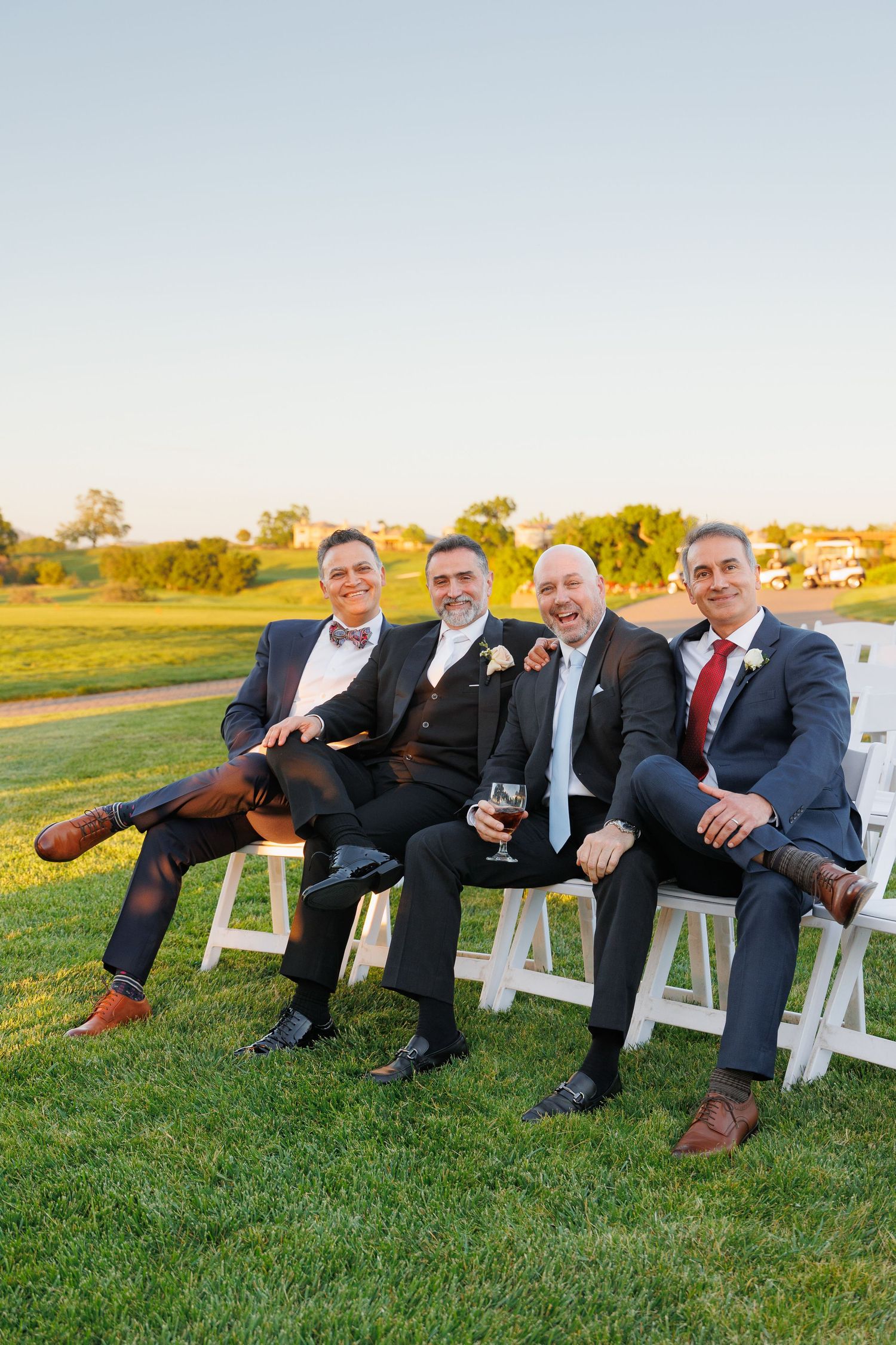 Four men in formal suits sitting on white chairs on a grassy lawn during golden hour.