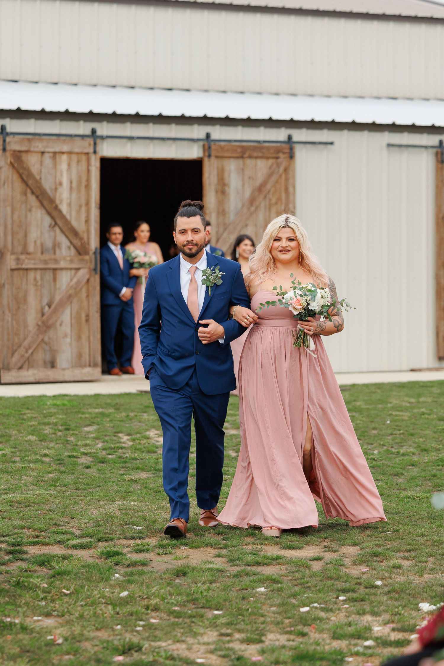 Wedding party members in navy suits and pink dresses walk and laugh together on grass outside a rustic barn venue.