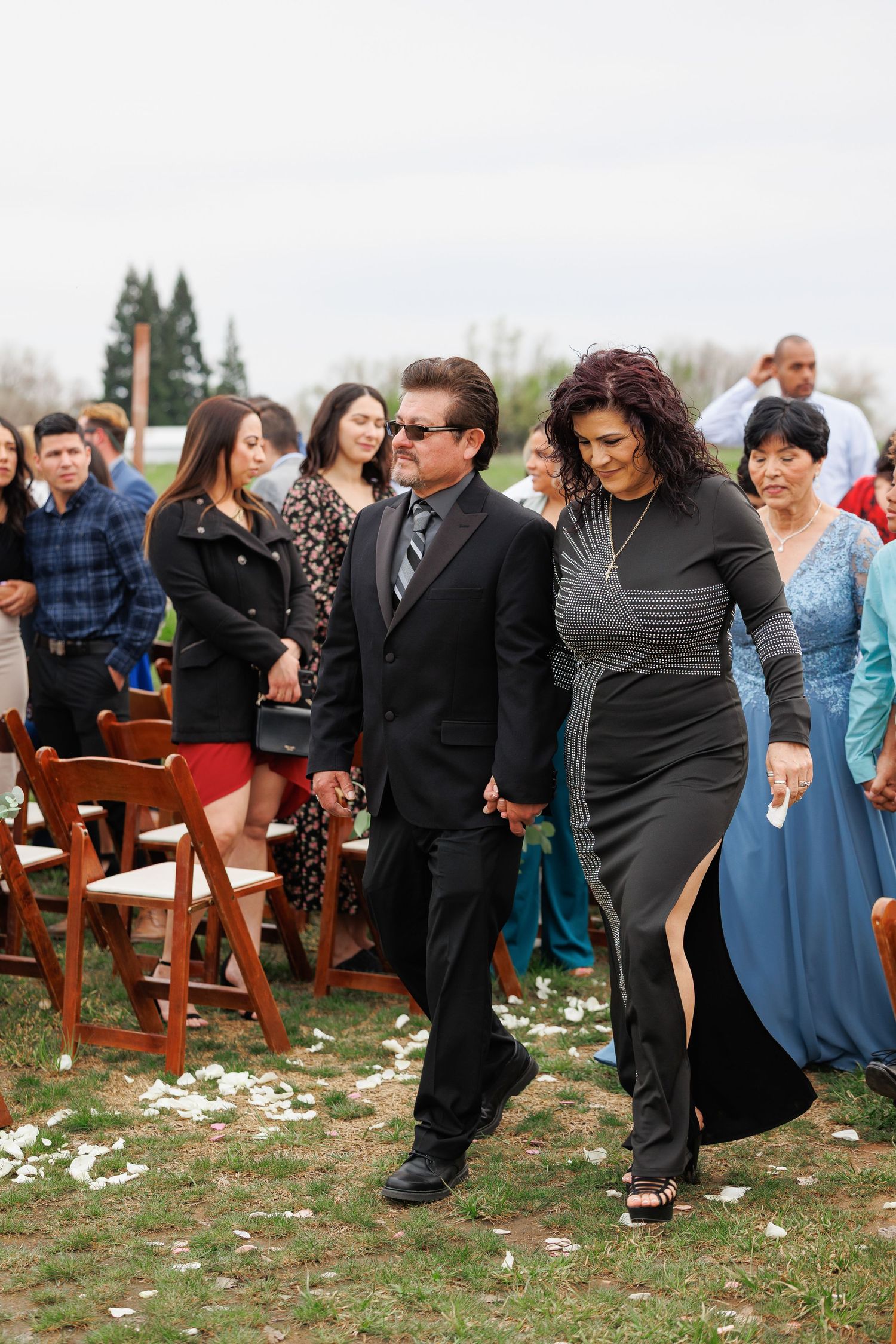 Wedding guests dance and celebrate together on a grassy outdoor venue with wooden chairs and scattered flower petals.