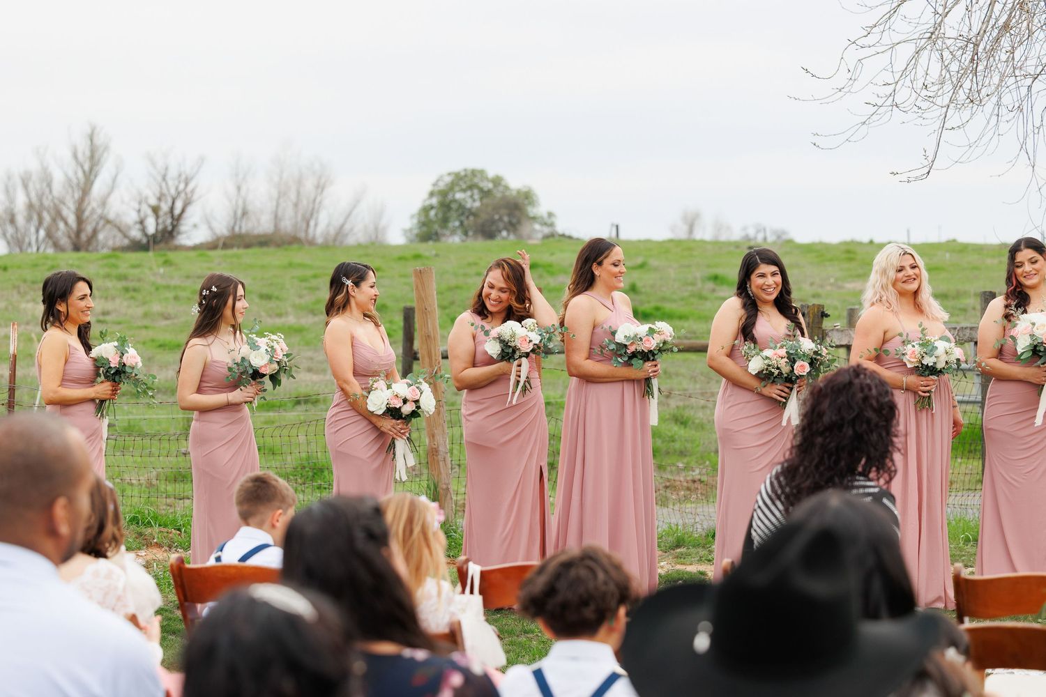 A wedding party in blush pink bridesmaid dresses poses outdoors on a green hillside during a spring ceremony.