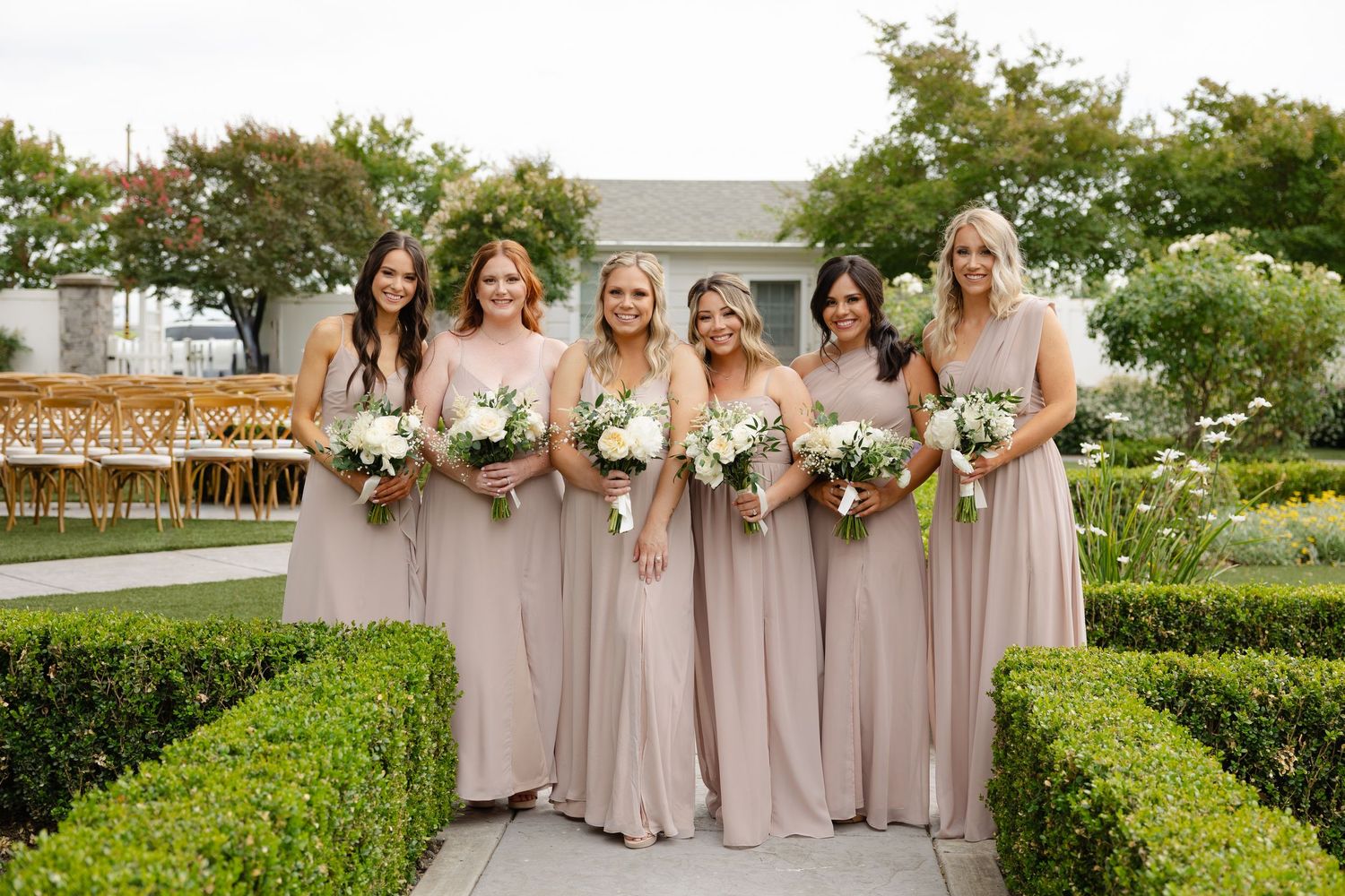 A group of bridesmaids in flowing champagne-colored dresses pose with white bouquets in a manicured garden setting.