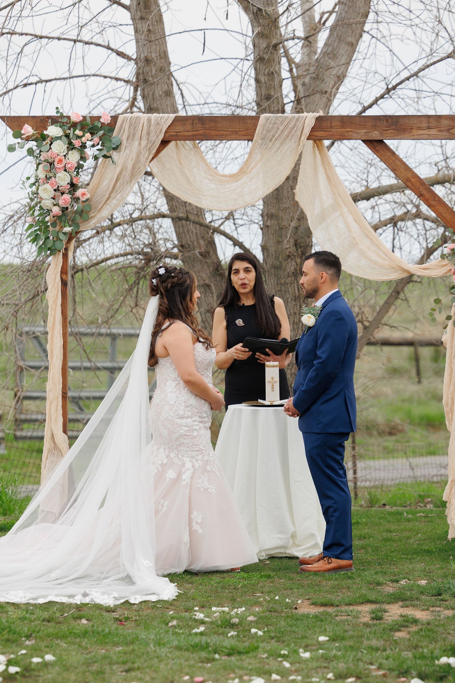 Wedding ceremony under wooden arch with white drapes and floral decorations in natural outdoor setting.