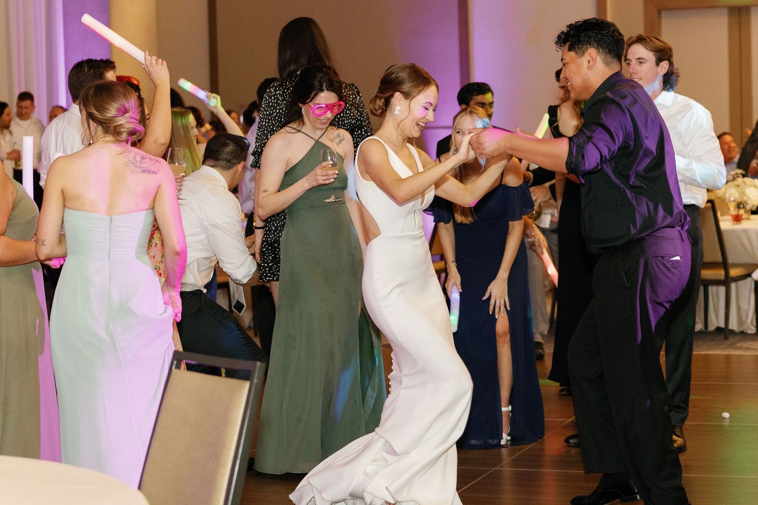 Wedding guests dance and celebrate on a ballroom dance floor illuminated by pink and purple lighting.
