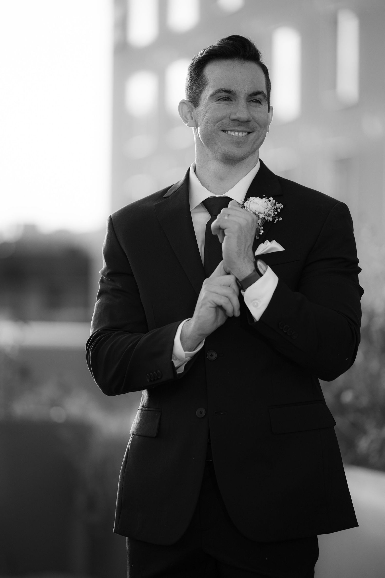 A smiling groom in a black suit with boutonniere adjusts his tie in this black and white wedding portrait.