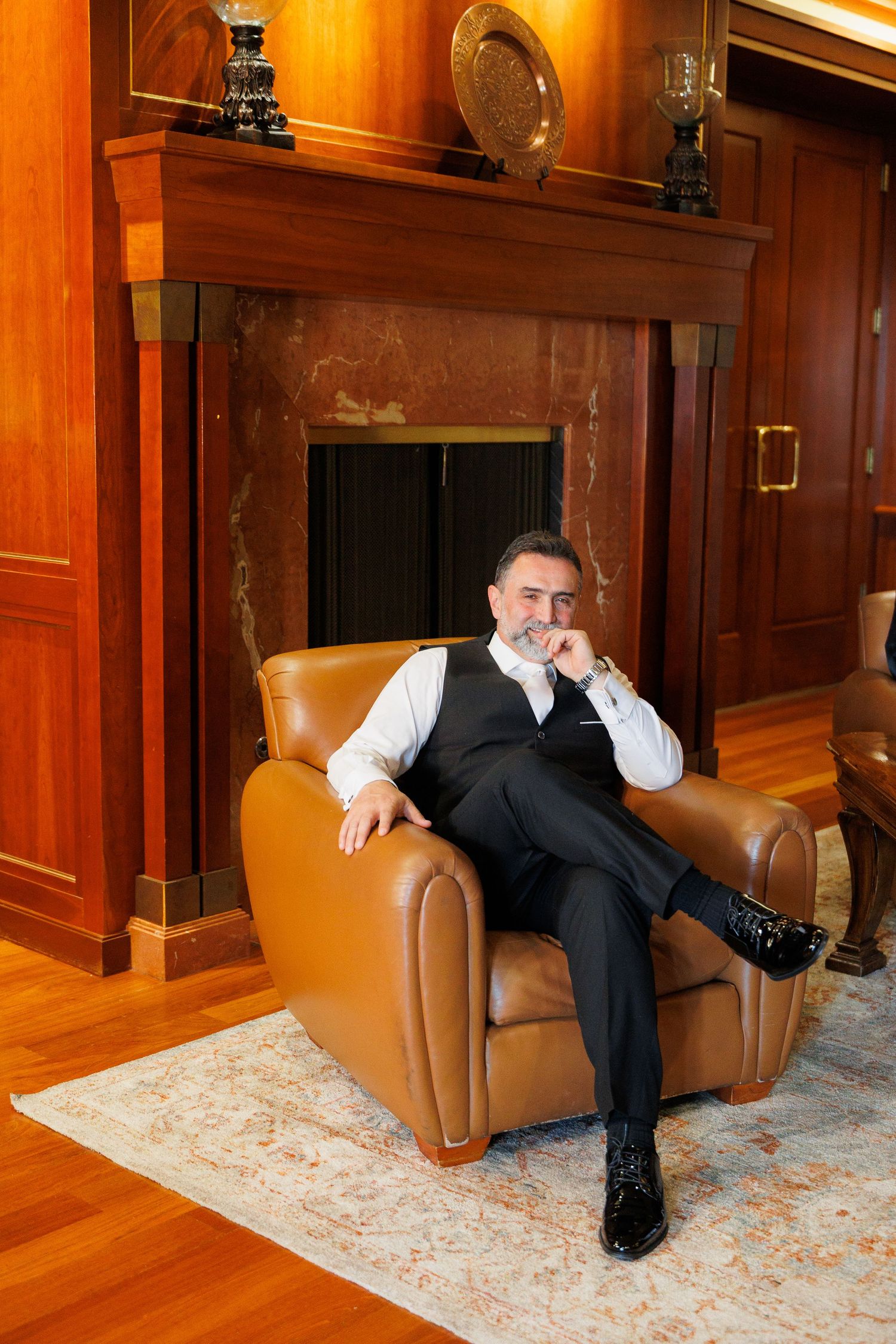 A person sits on a brown leather chair in an elegant wood-paneled room with a decorative area rug.